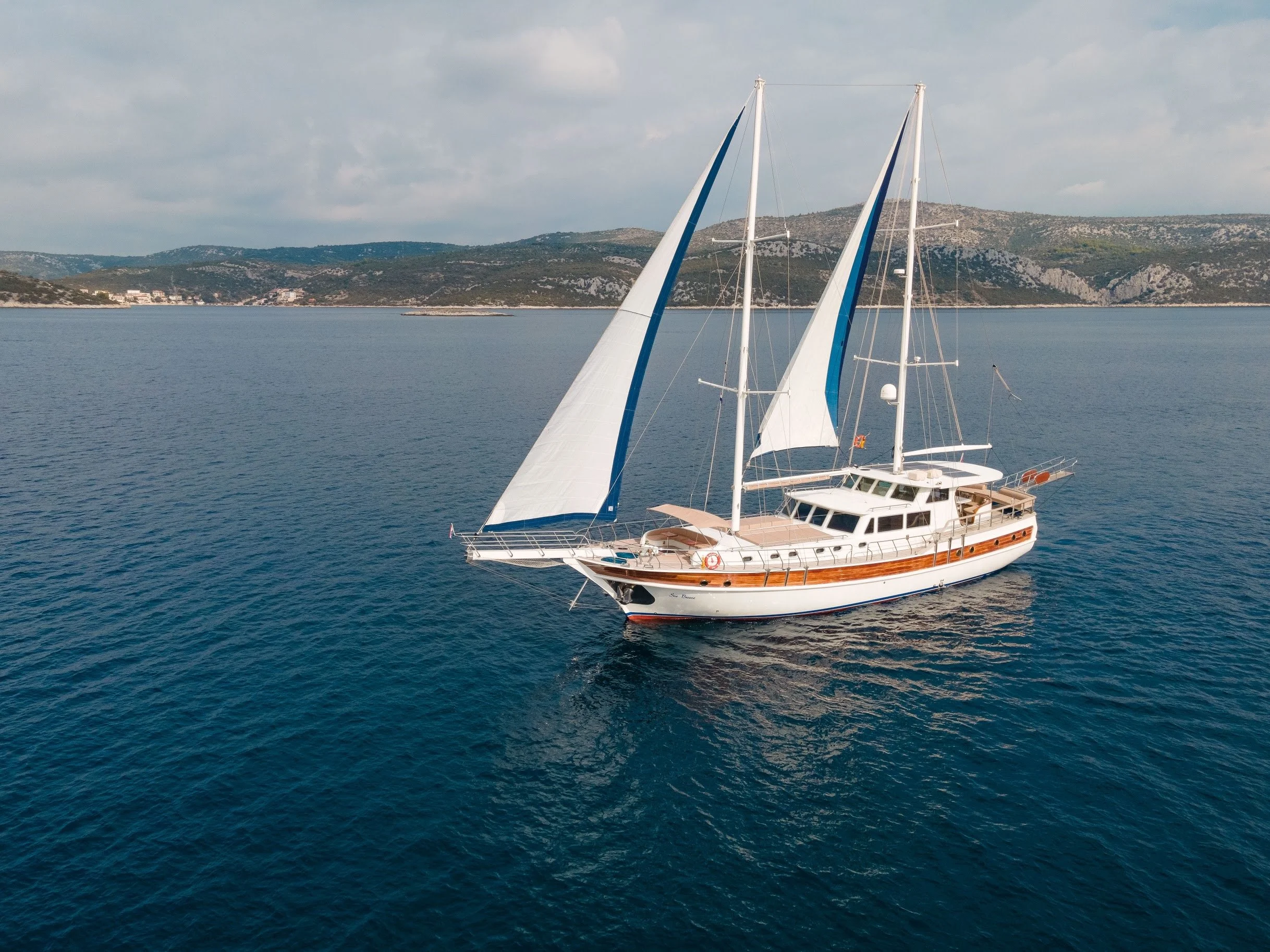 A sailboat with two masts and white sails on calm blue water near a hilly coastline.