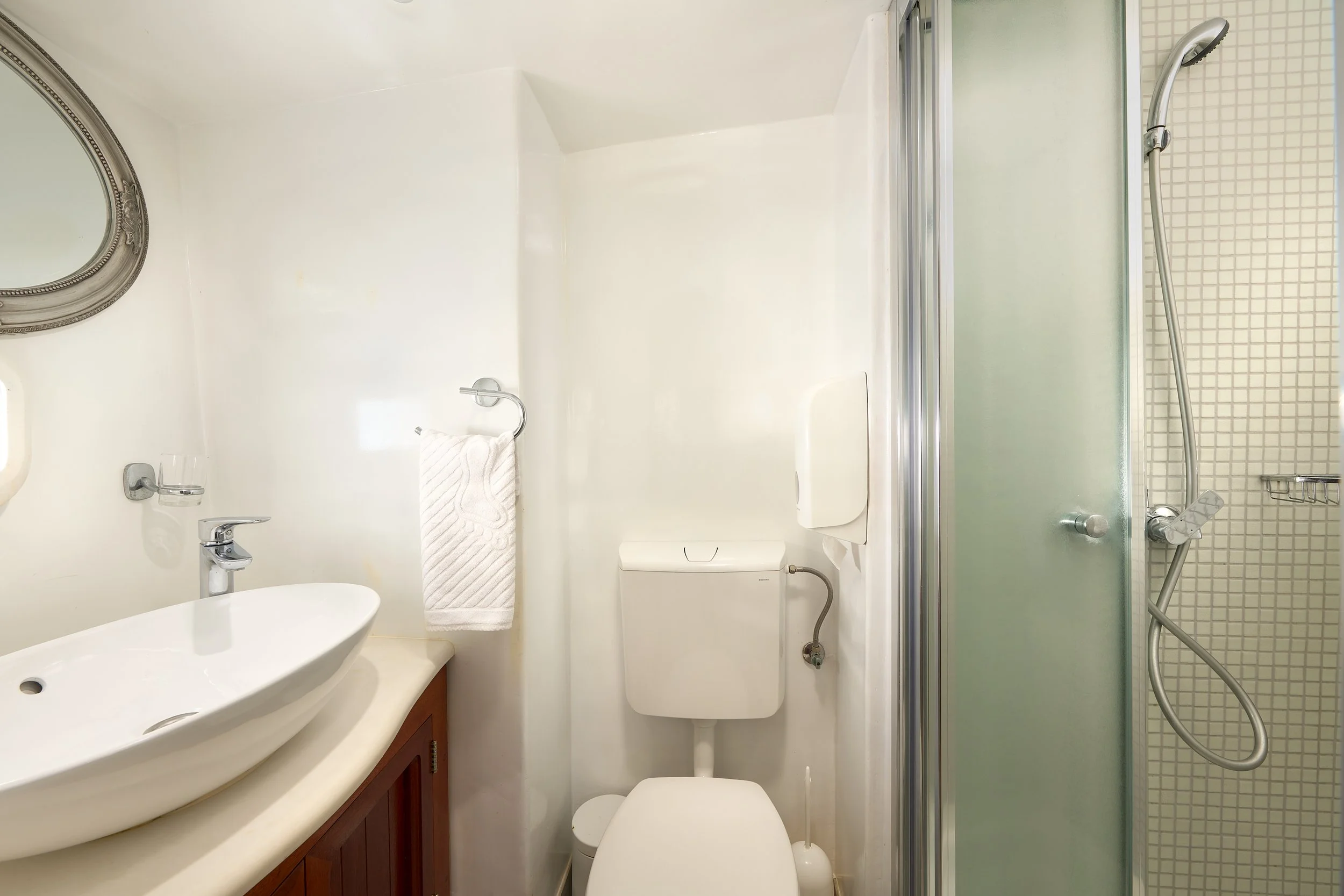 Small bathroom with a white sink, round mirror, toilet, a small towel on a hook, and a glass shower enclosure with a handheld showerhead.