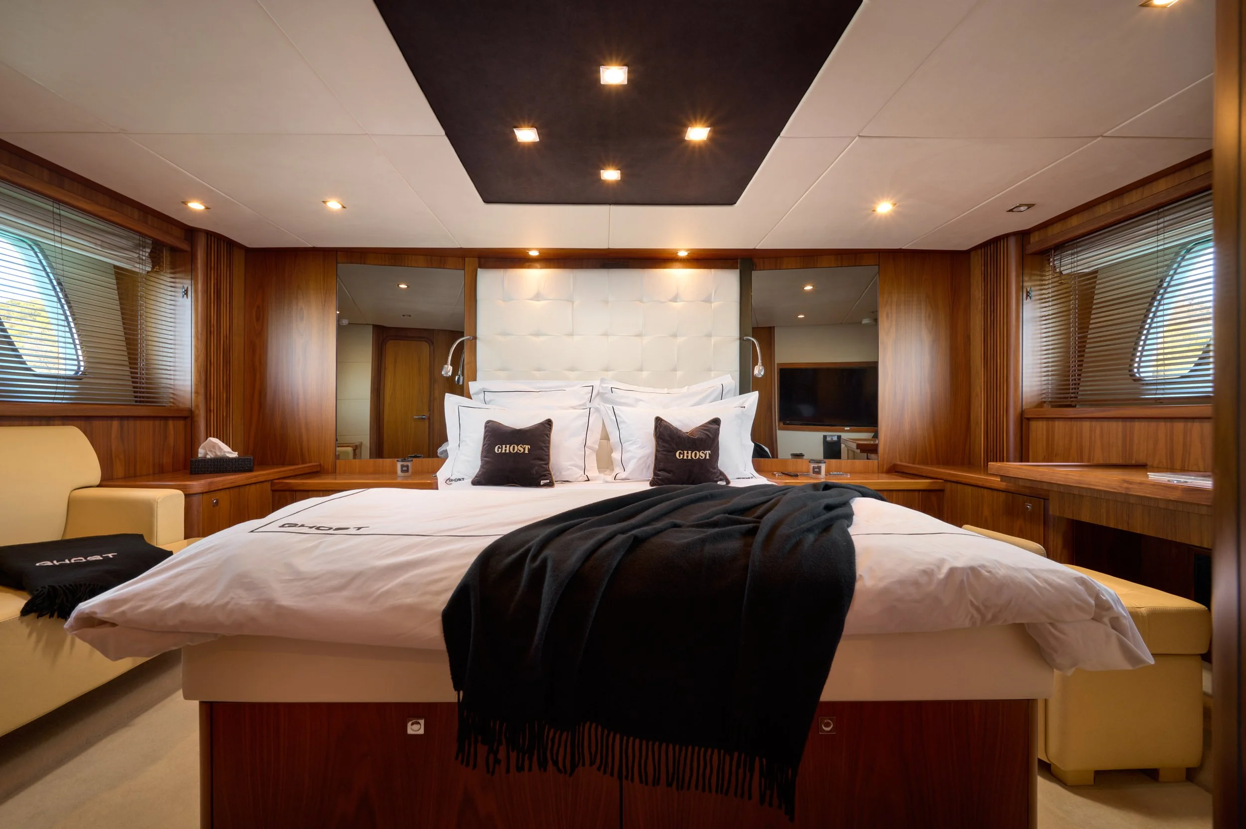 Luxurious bedroom with wooden walls, a large bed with white bedding, and black pillows with 'GHOST' written on them. The room features a white padded headboard, modern wall-mounted reading lamps, a flat-screen TV, and large windows with blinds. A bla