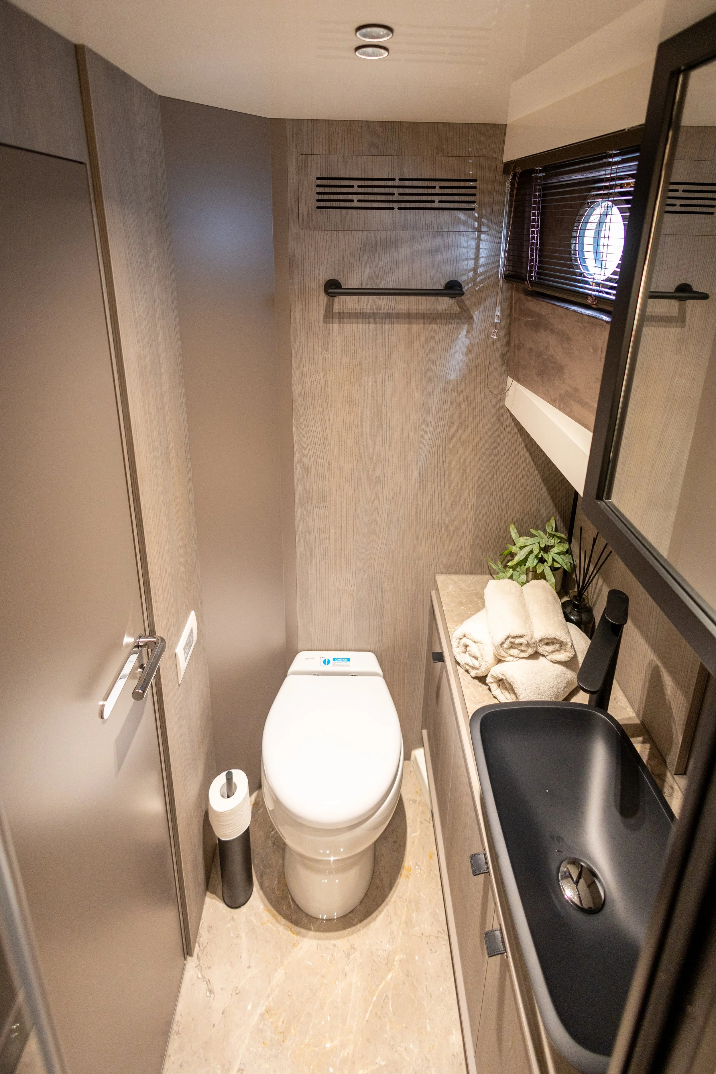 Small modern bathroom with a toilet, rolled towels, a black sink, and a small window with blinds.