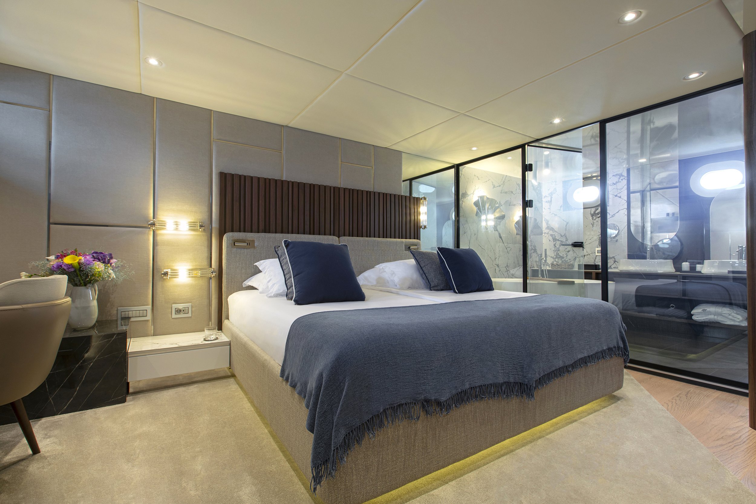 A modern hotel bedroom with a bed featuring blue pillows and a blue throw blanket, a side table with flowers, and a glass-enclosed bathroom in the background.