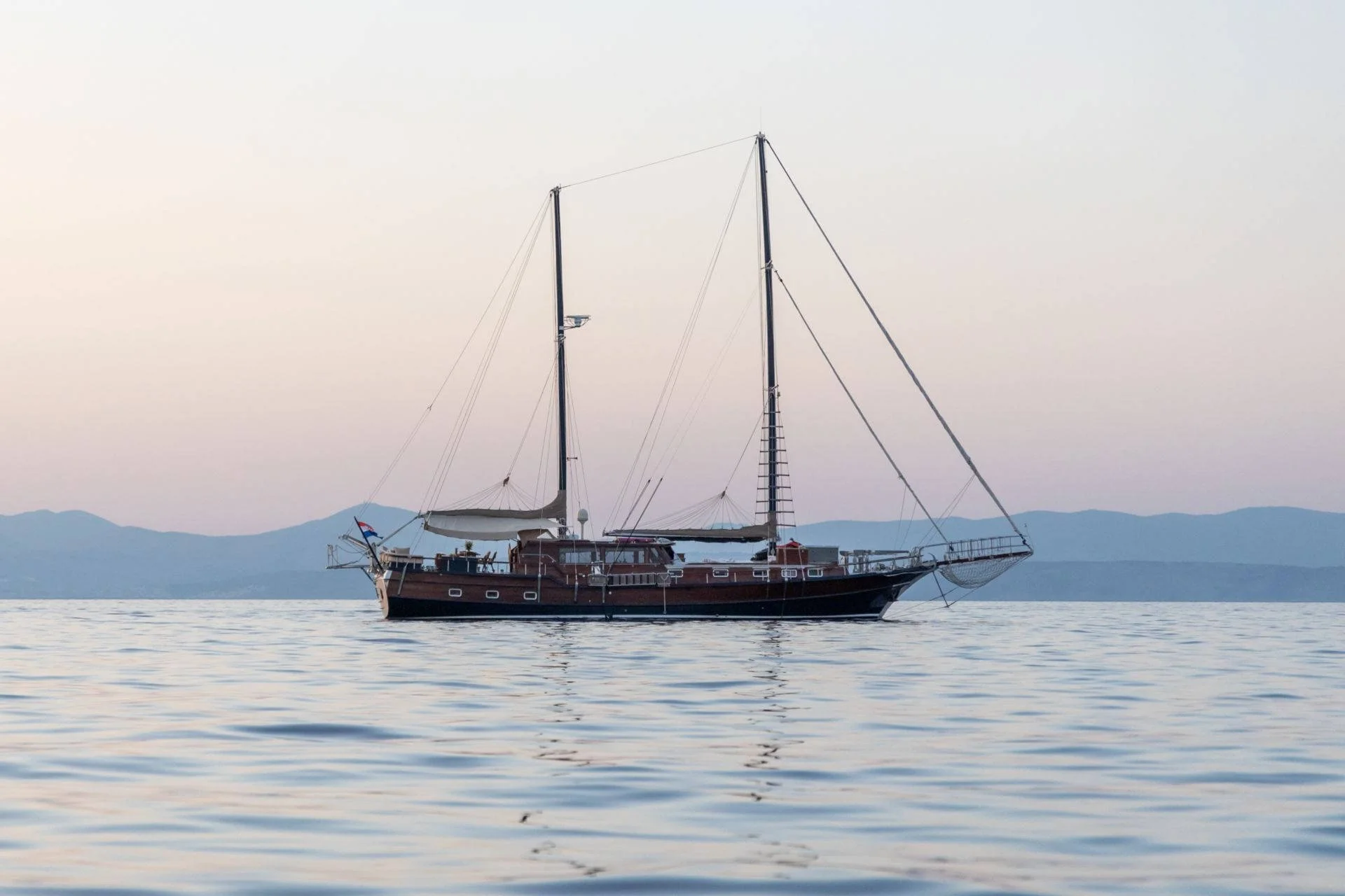 Sailing ship on calm sea at sunset with mountains in background.