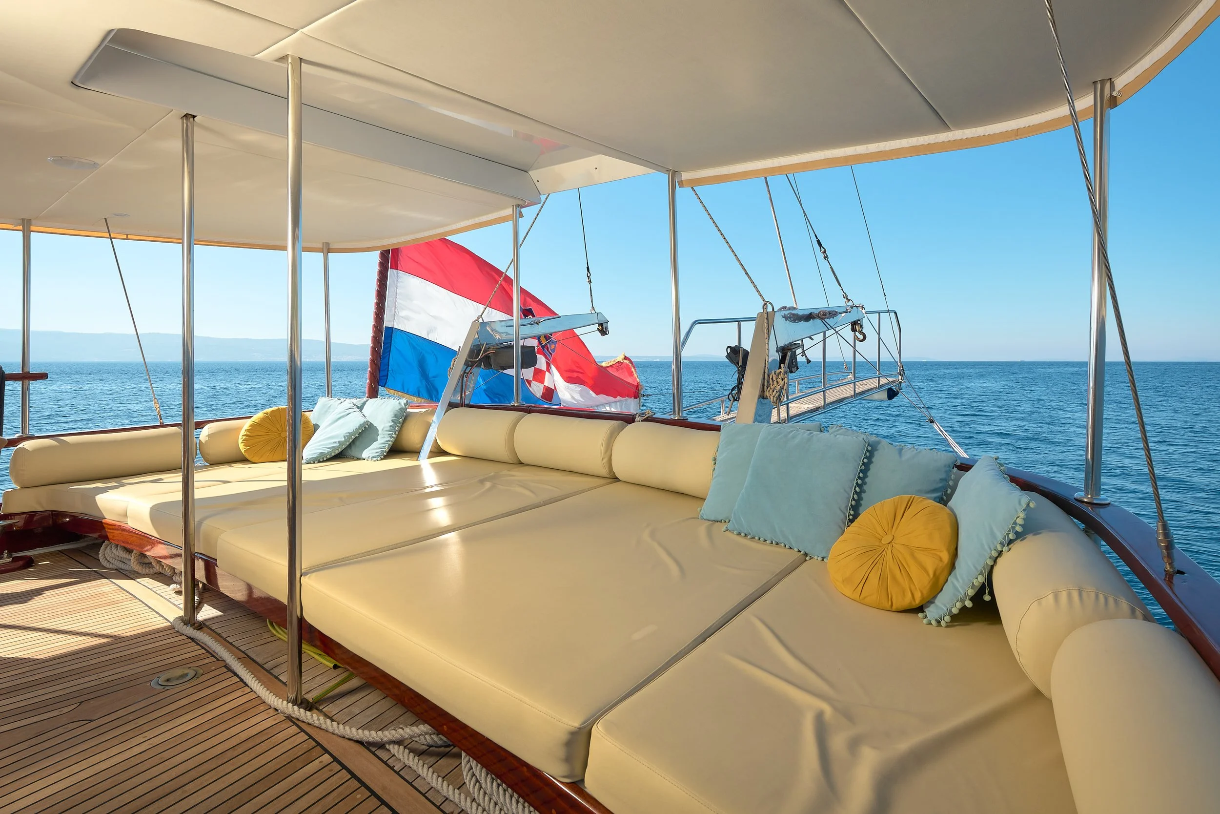 View of a yacht deck with yellow cushioned seating, blue and yellow pillows, a Croatian flag, and a clear ocean view in the background.