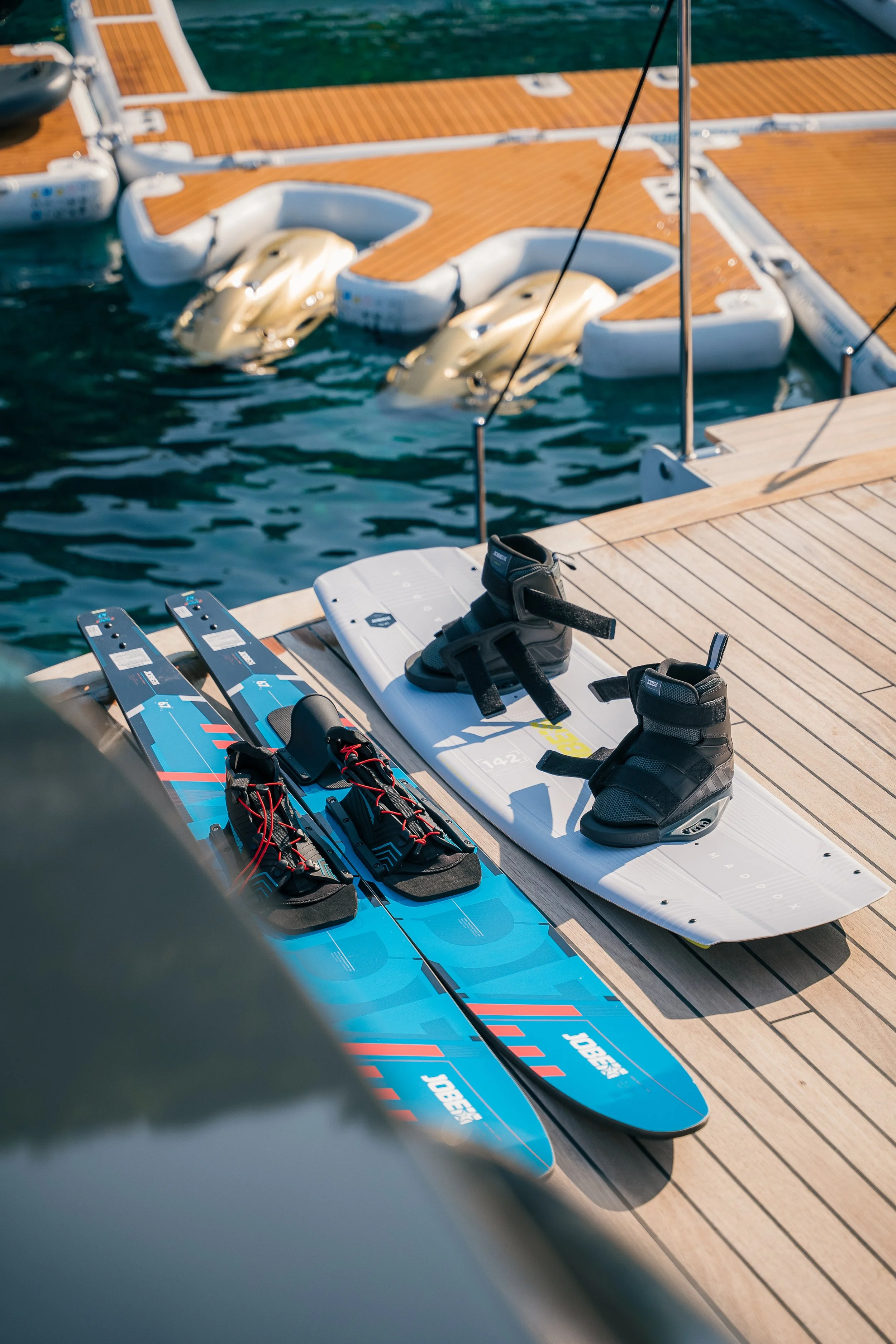 Water skis and a wakeboard with bindings on a wooden dock by the water, with pedal boats in the background.