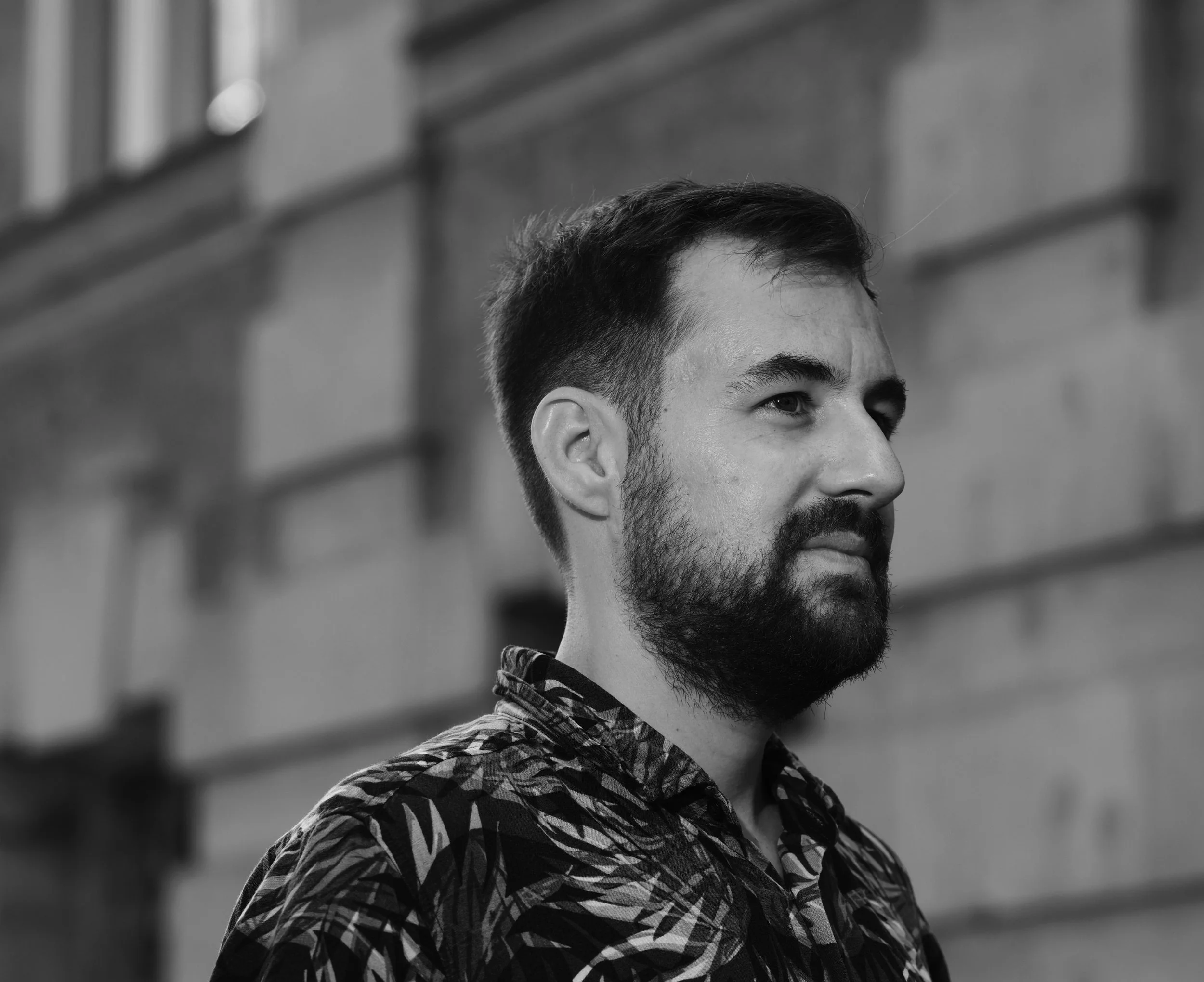 Black and white photo of a man with a beard and short hair, wearing a patterned shirt, standing outdoors against a brick wall.