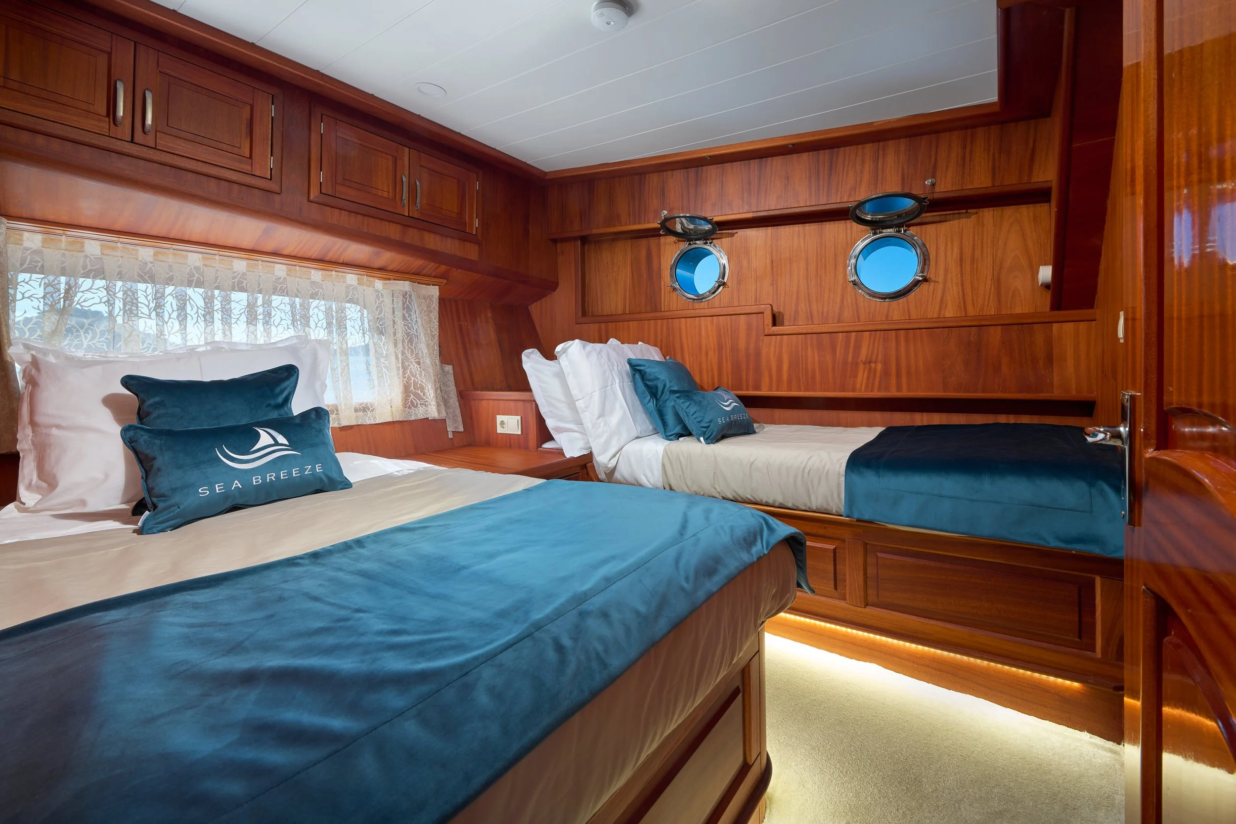 Two beds with Sea Breeze pillows in a wooden-paneled bedroom with small porthole windows.