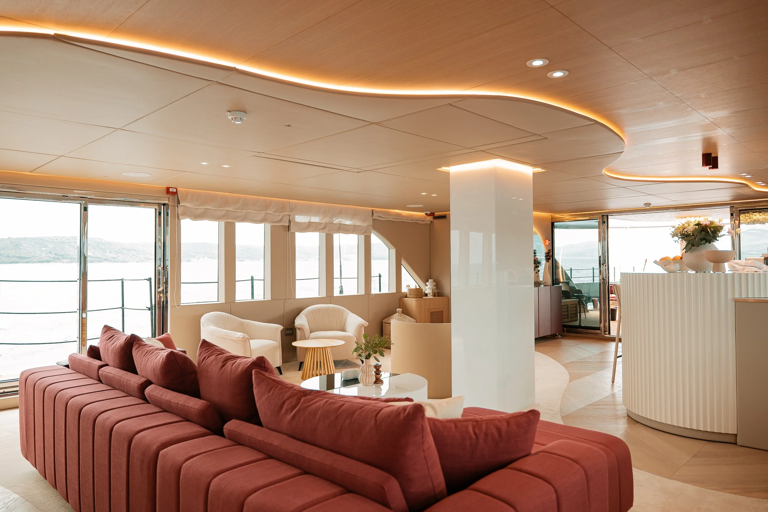 Interior of a modern, well-lit lounge area on a cruise ship with a large red sofa, white armchairs, and windows overlooking water and landscape.