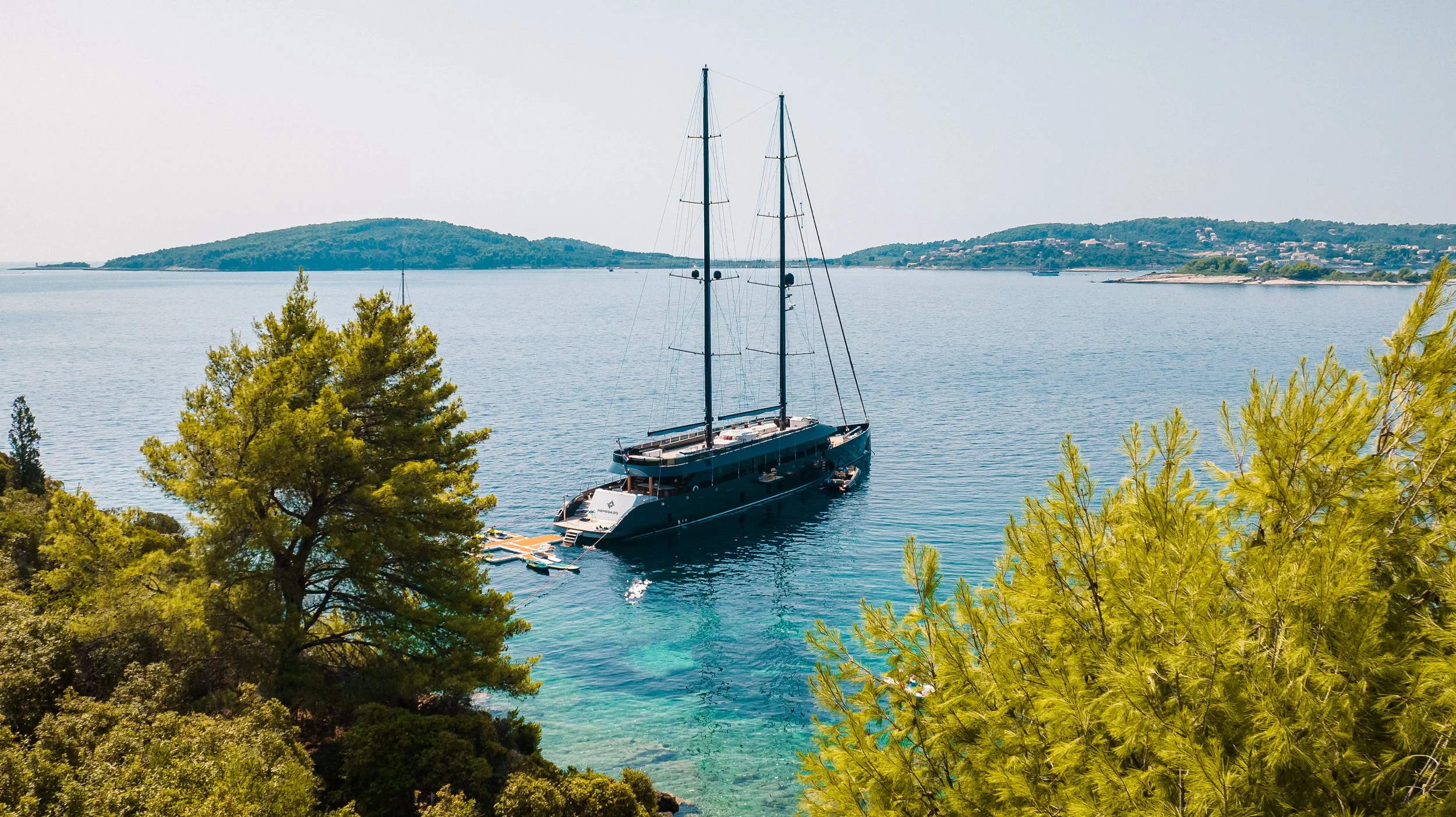 A large black sailboat is anchored in a serene, bright blue body of water, surrounded by lush green trees on the shoreline, with distant hills on the horizon.