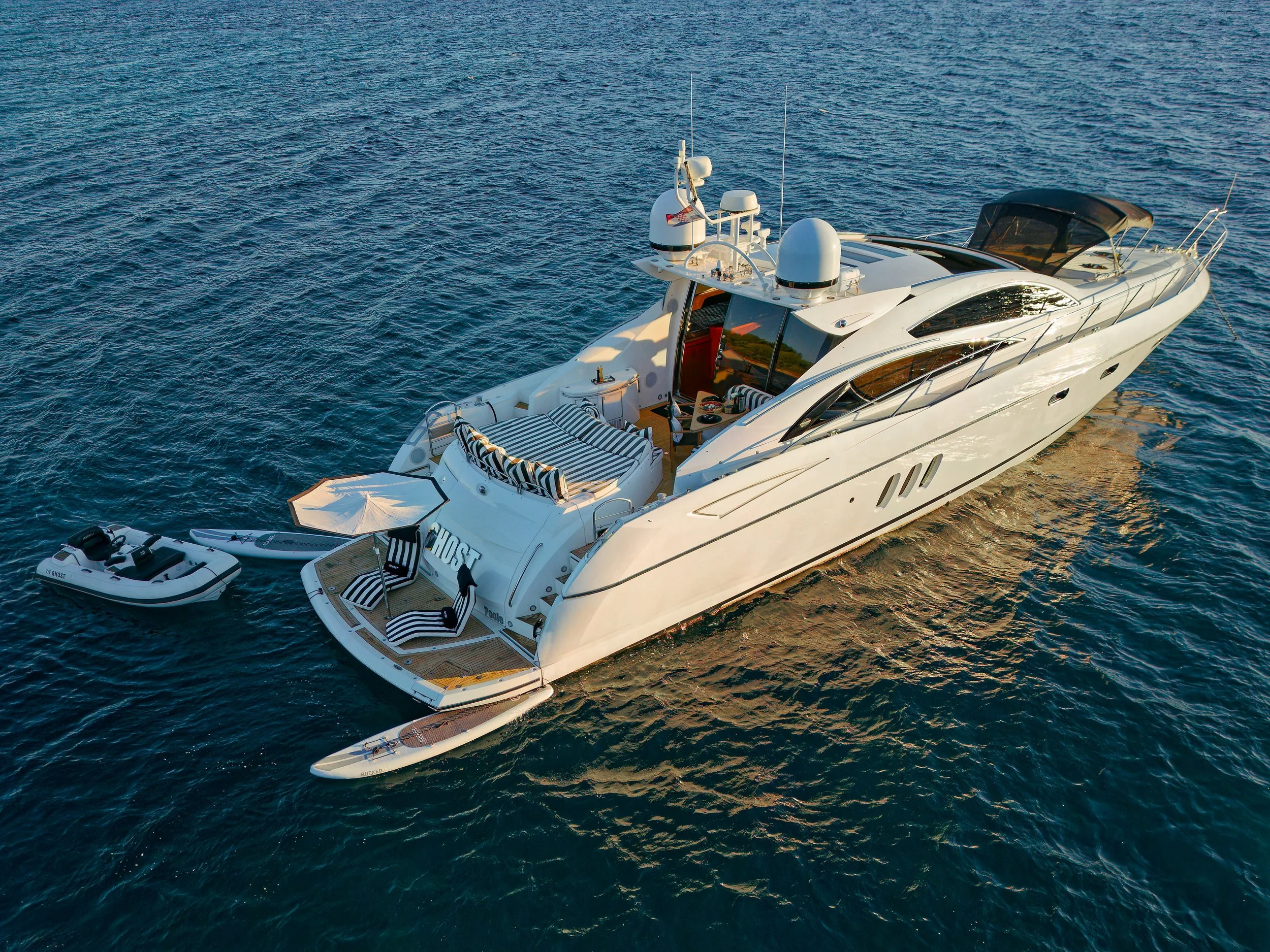 Aerial view of a luxury white yacht on the ocean with a small inflatable boat tied to its side, featuring a sun umbrella and striped seating.