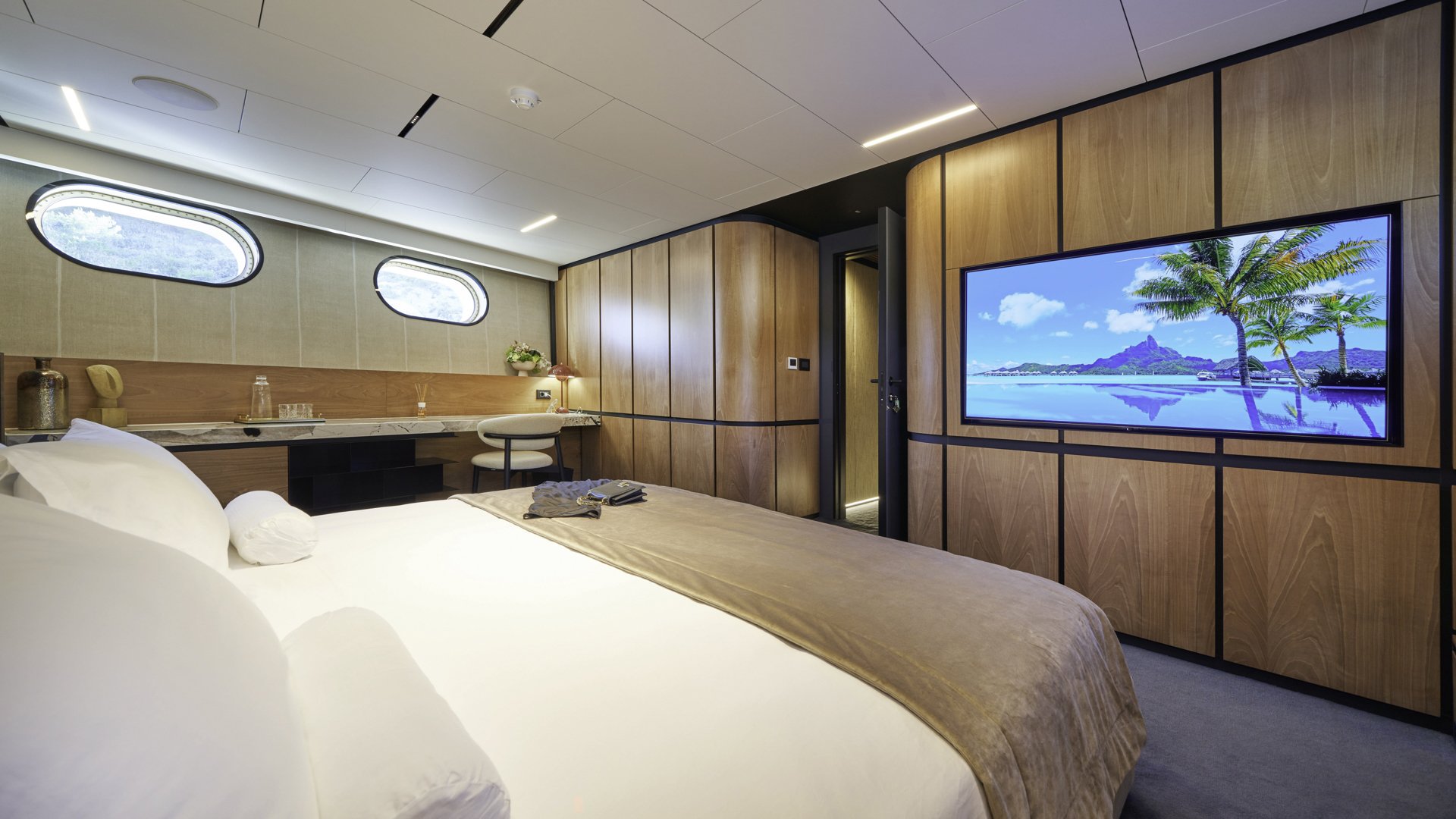 Modern hotel room with a large bed, a wall-mounted TV displaying a tropical beach scene with palm trees and mountains, wooden paneling on the walls, and small oval windows letting in natural light.