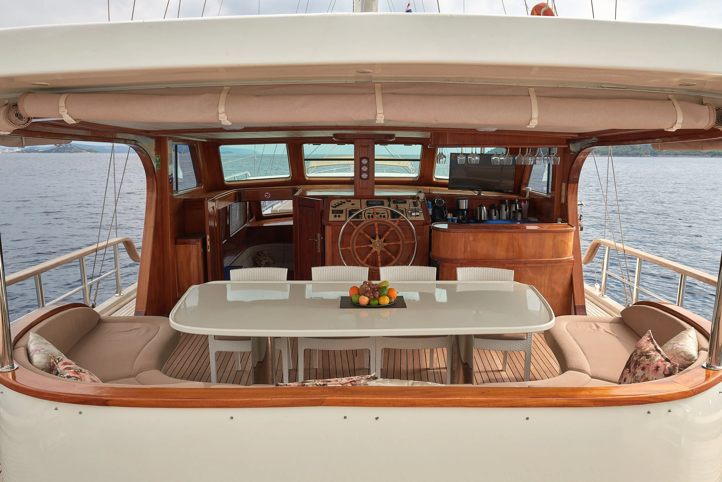 Interior view of a luxury yacht's deck showing a white dining table with six chairs, floral pillows on bench seating, a fruit centerpiece, and the boat's wooden helm station with steering wheel and navigational equipment.