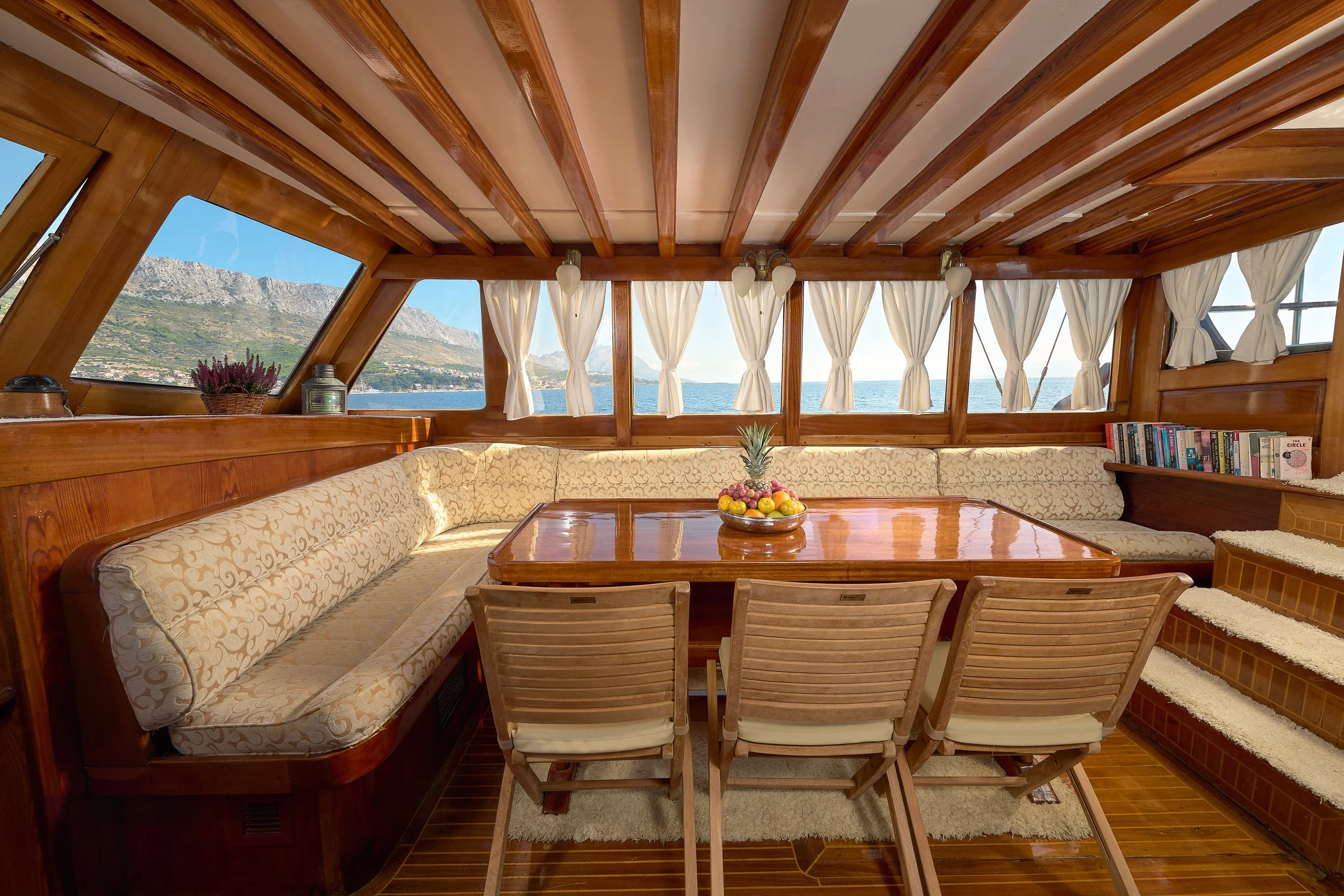 Interior of a boat with wooden walls and ceiling, a U-shaped cushioned seating area, a wooden table with a bowl of fruit, and large windows showing a view of the water and distant mountains.
