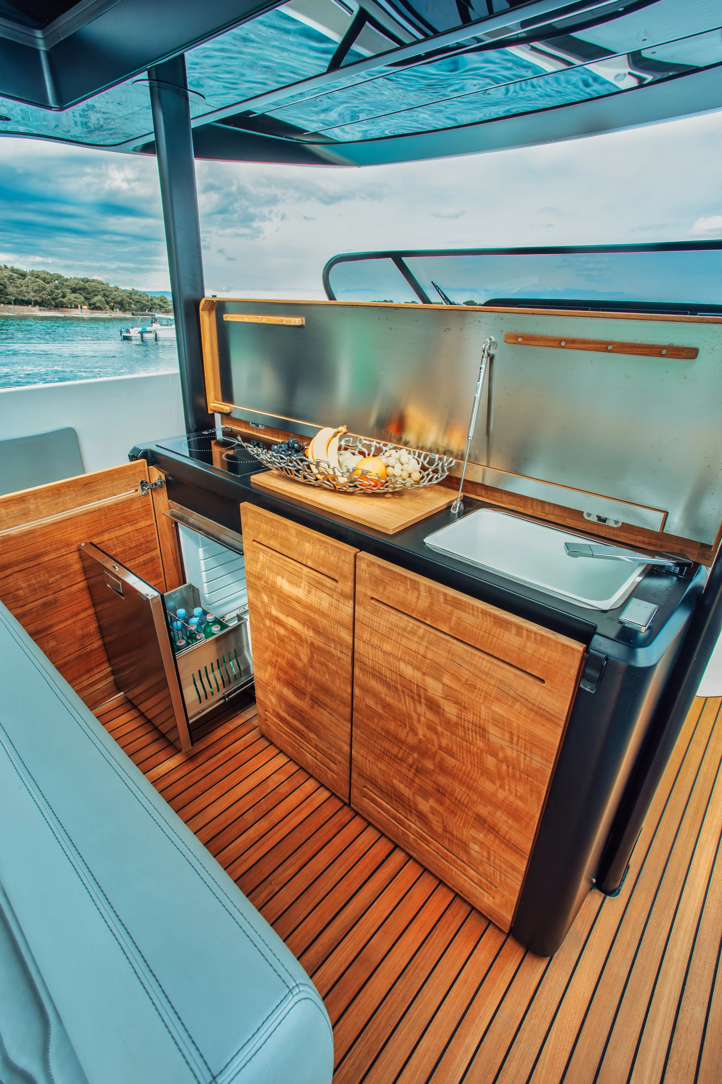 A boat's outdoor kitchen area with a wooden cabinet, small fridge, and a countertop with a sink and fruit on it, surrounded by water and sky.