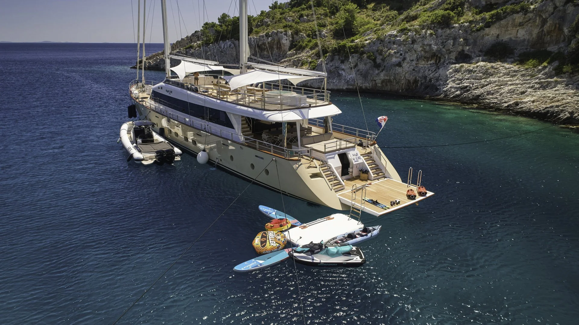 A luxury yacht anchored near a rocky green coastline, with smaller boats and paddleboards around it.