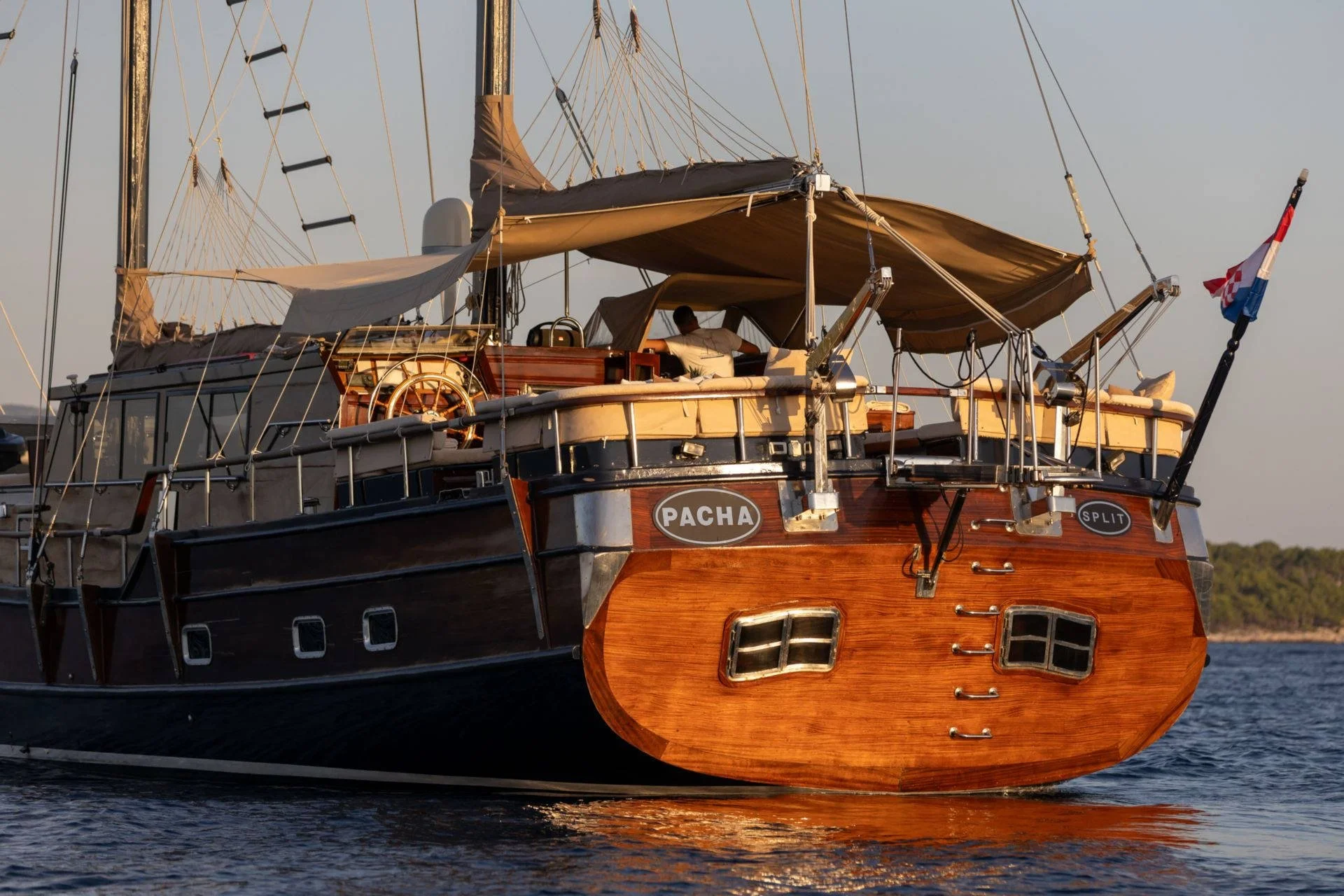 Wooden yacht with sails, named "Pacha," on water at sunset.