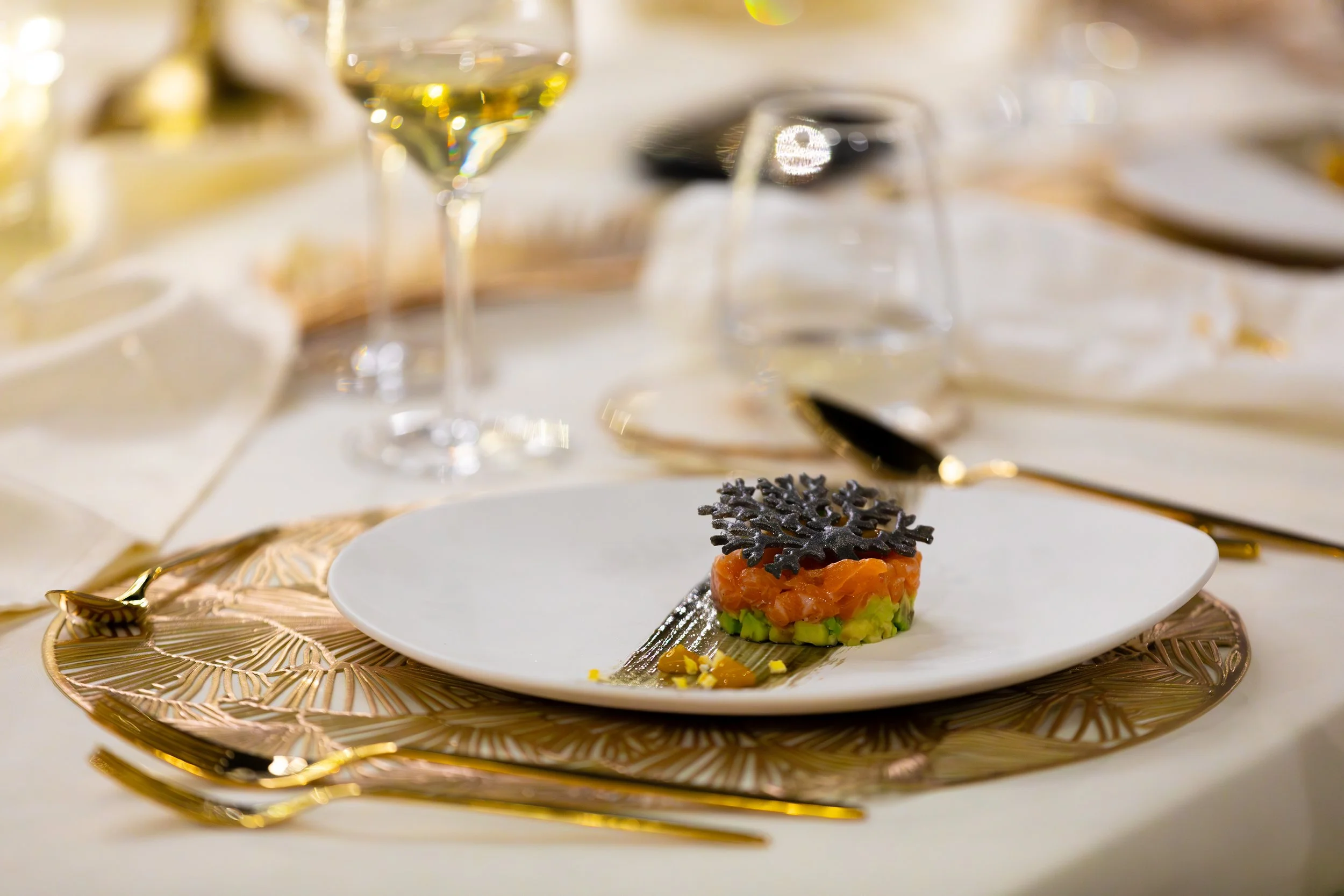Elegant dining table with a white plate holding a small serving of salmon tartare topped with black garnish, gold and black cutlery, wine glasses, and a decorative charger plate.