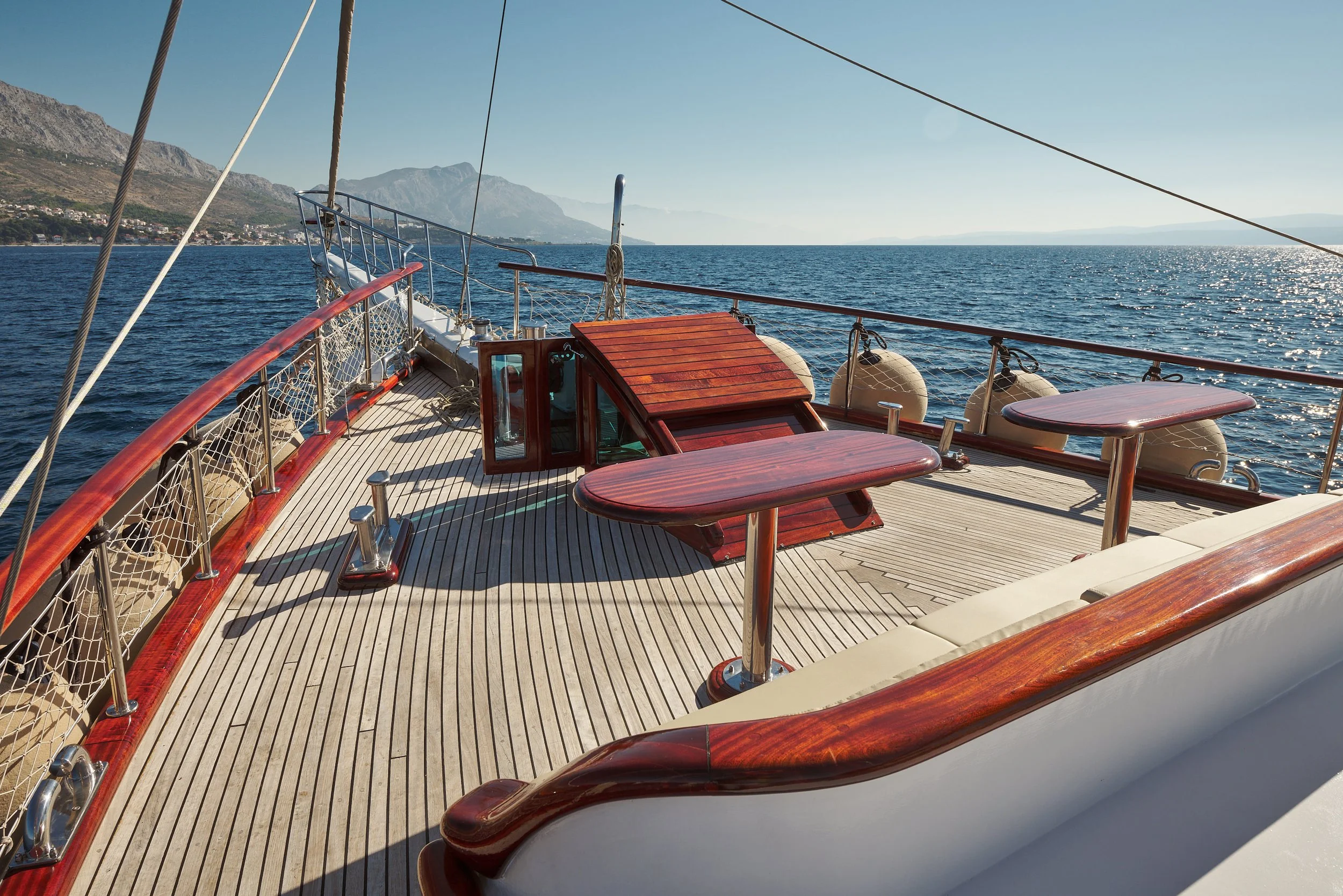 View of the deck of a luxury yacht with wooden accents, tables, and safety equipment, overlooking the ocean with mountains in the distance under clear skies.