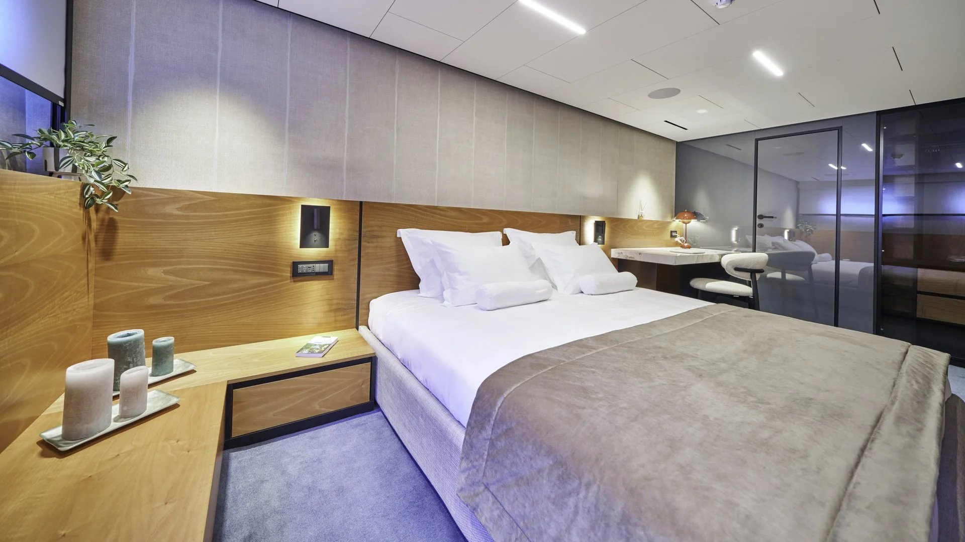Modern hotel bedroom with a king-sized bed, white linens, and wooden headboard, beside a nightstand with candles and a magazine, a glass sliding door reflecting the room, and a desk with a chair and reading lamp.