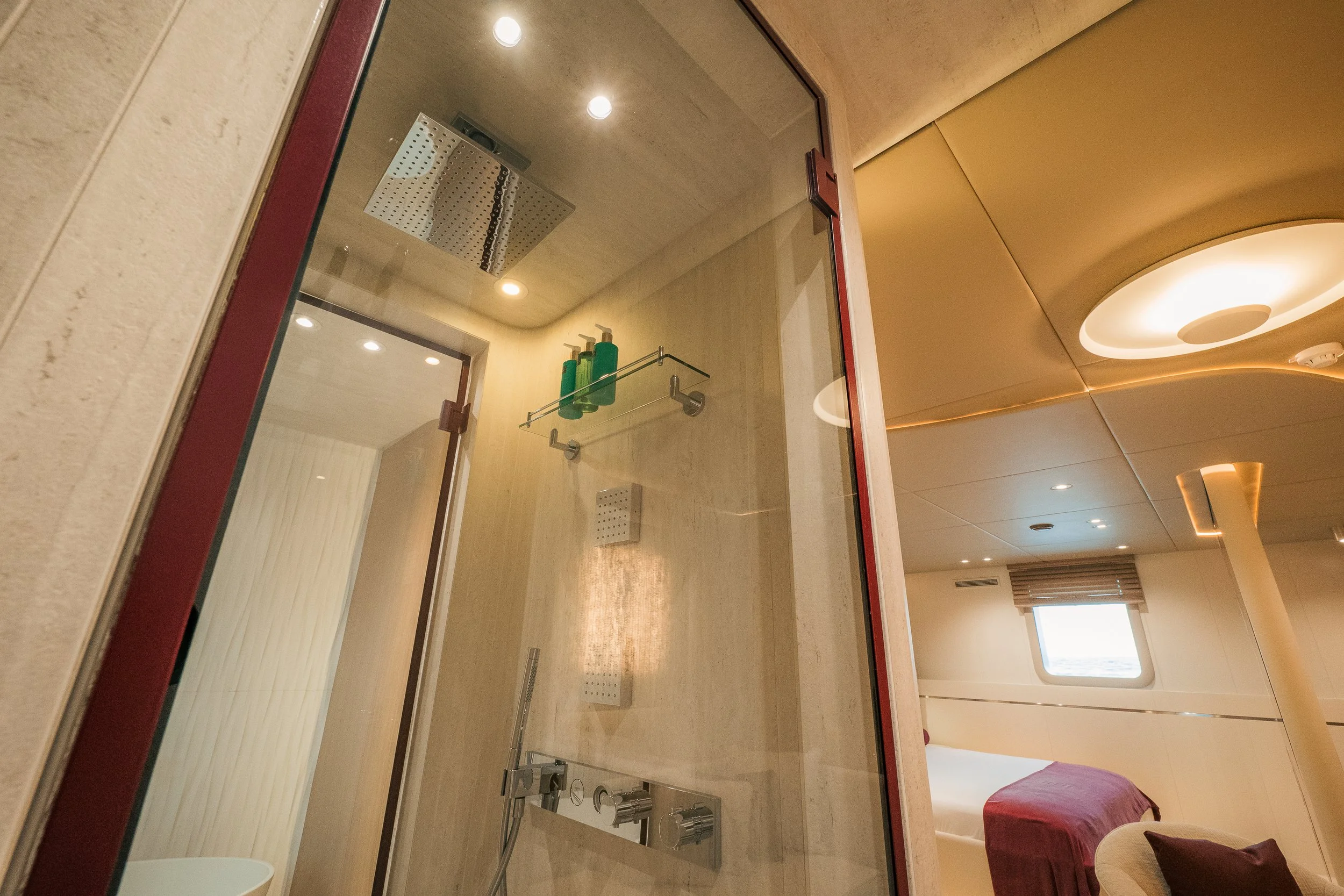 A modern glass-enclosed shower with a rainfall showerhead, mounted on a beige marble wall. Inside the shower, three green bottles are on a glass shelf. Adjacent to the shower is a bedroom with a bed dressed in dark red bedding, next to a small window