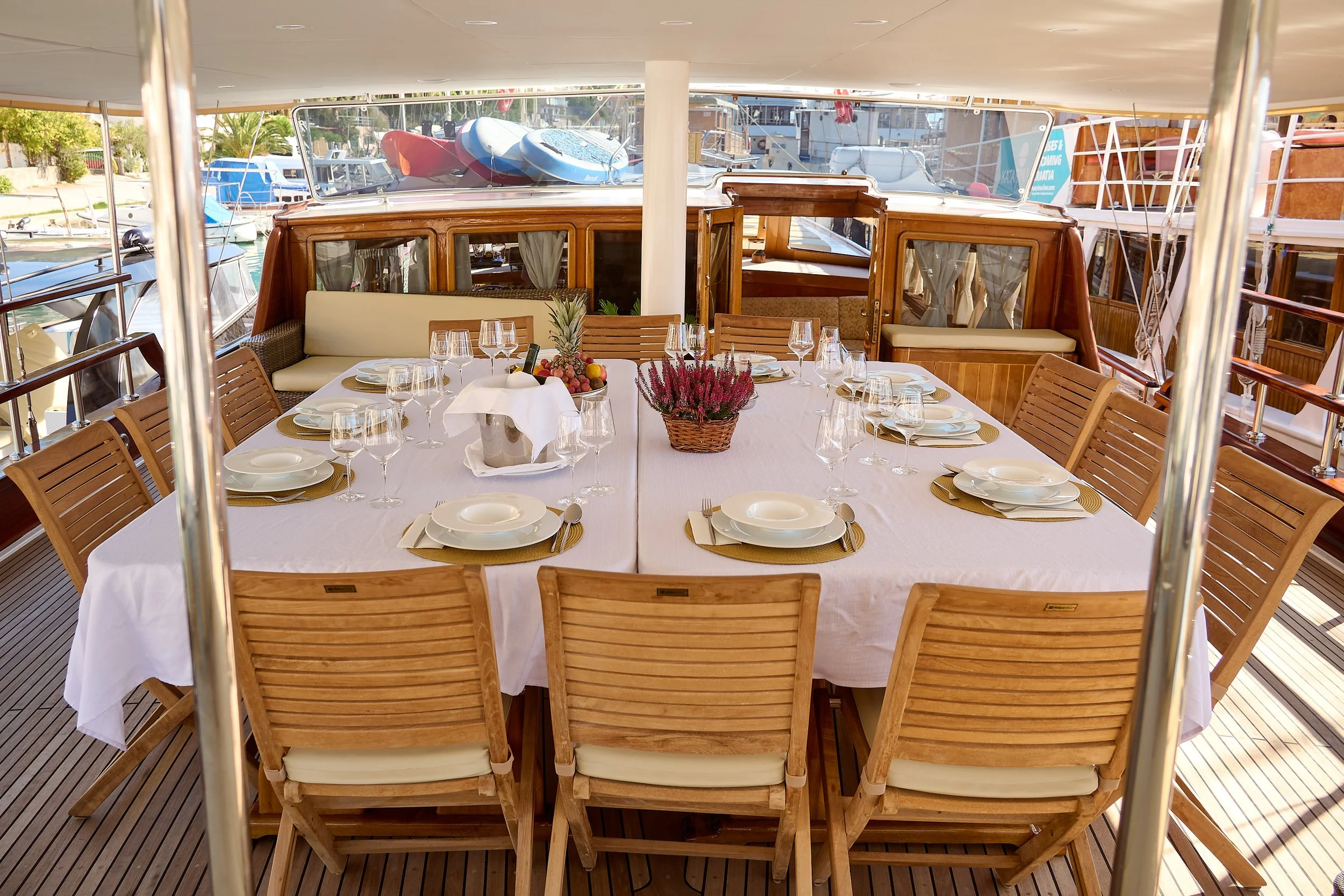 A dining table set with plates, glasses, and utensils on a yacht, with the marina and other boats visible in the background.