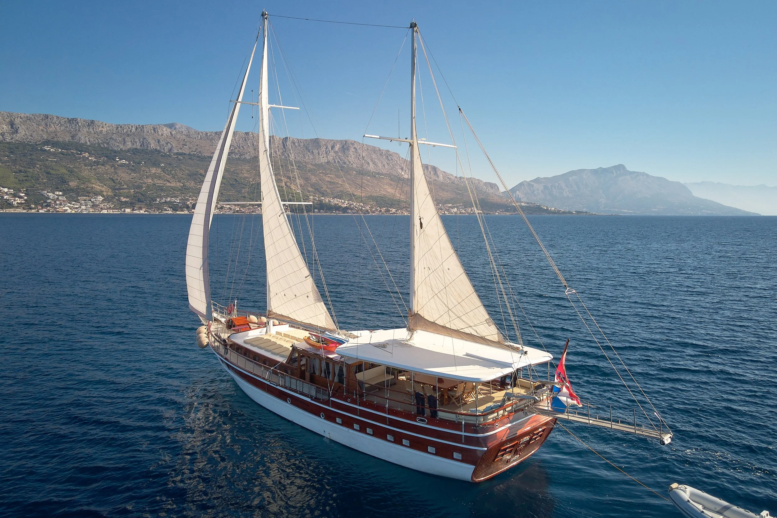 A large wooden sailboat with white sails on calm blue water, with a mountainous coastline in the background.