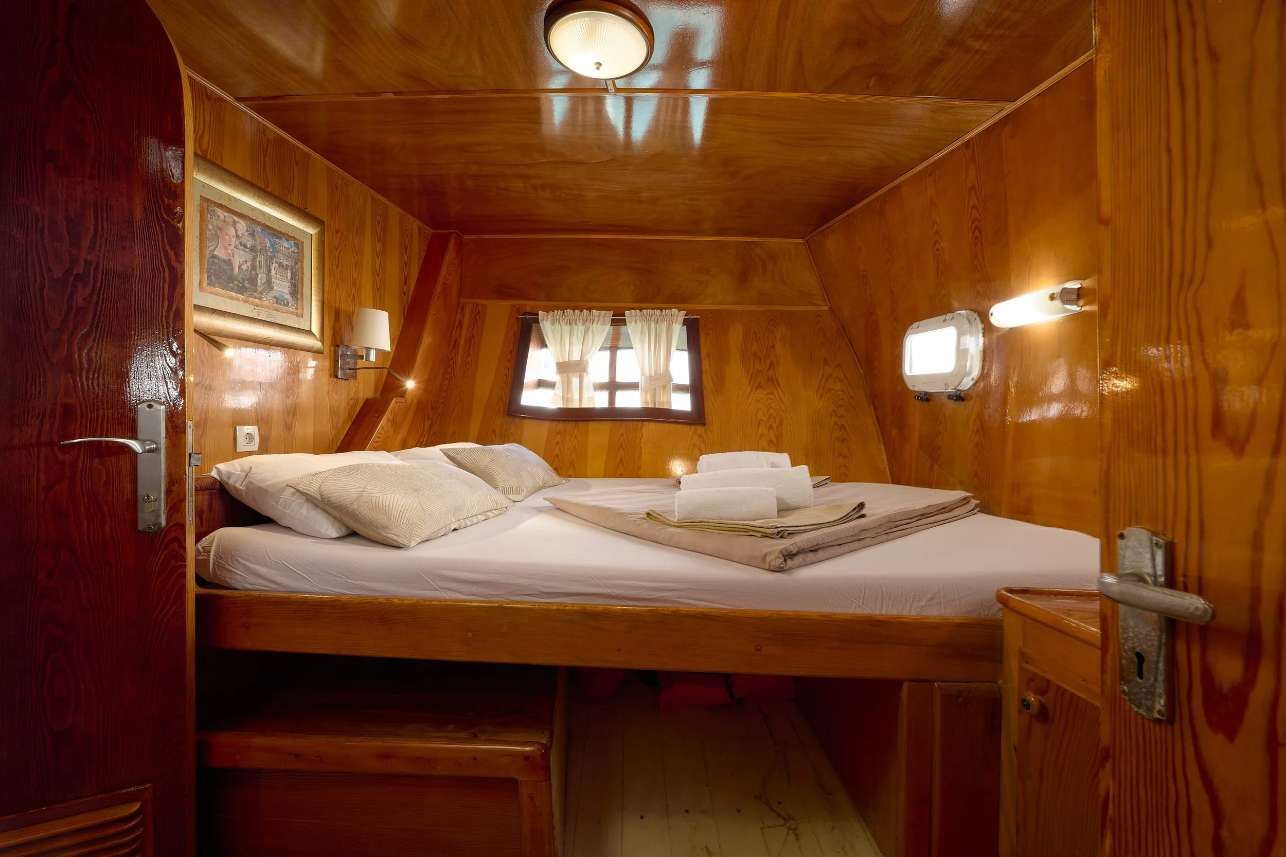 Cozy wooden interior bedroom with a bed, pillows, folded towels, small window with curtains, and wall-mounted lights.