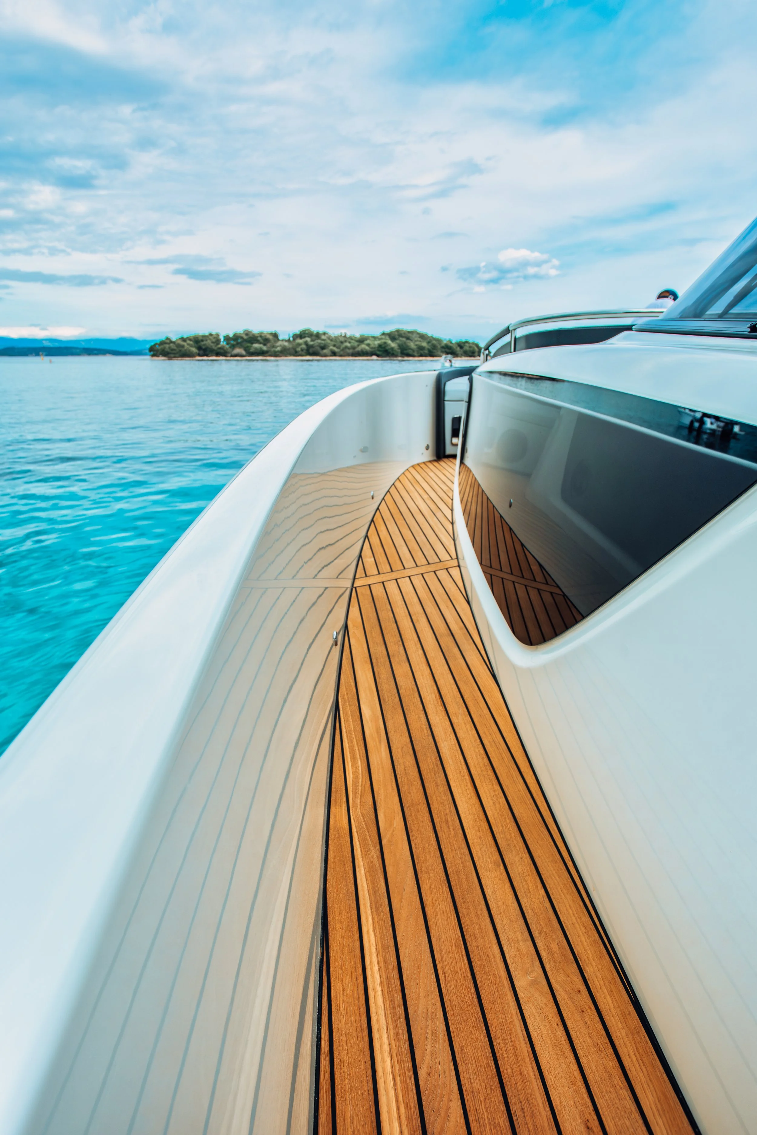 View from the side of a luxury yacht with a wooden deck, floating on blue water near a landmass with trees under a partly cloudy sky.