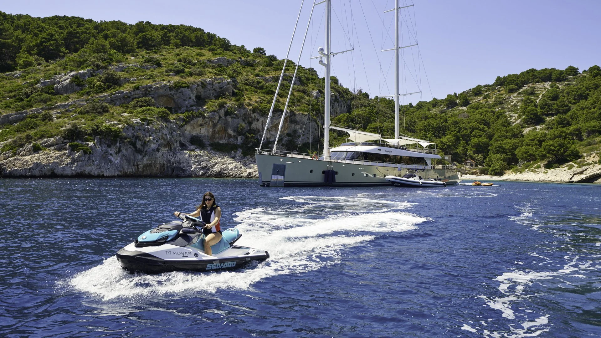 A woman riding a jet ski on the water near a large sailing yacht anchored in a cove with rocky, green hills in the background.