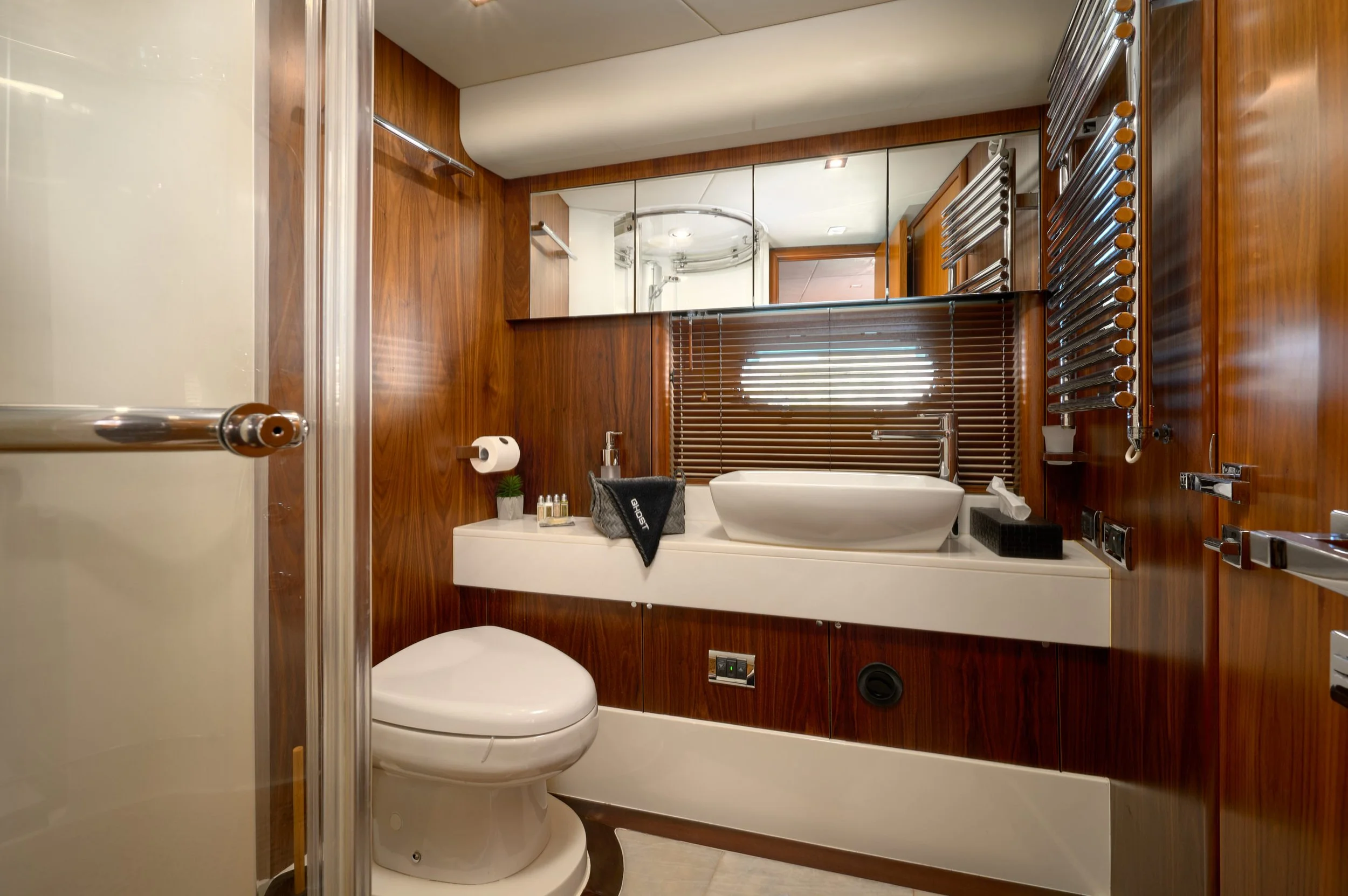Modern bathroom with wood-paneled walls, a white sink with a rectangular basin, a toilet, a large mirror, and wooden blinds on a window, with various toiletries and fixtures.