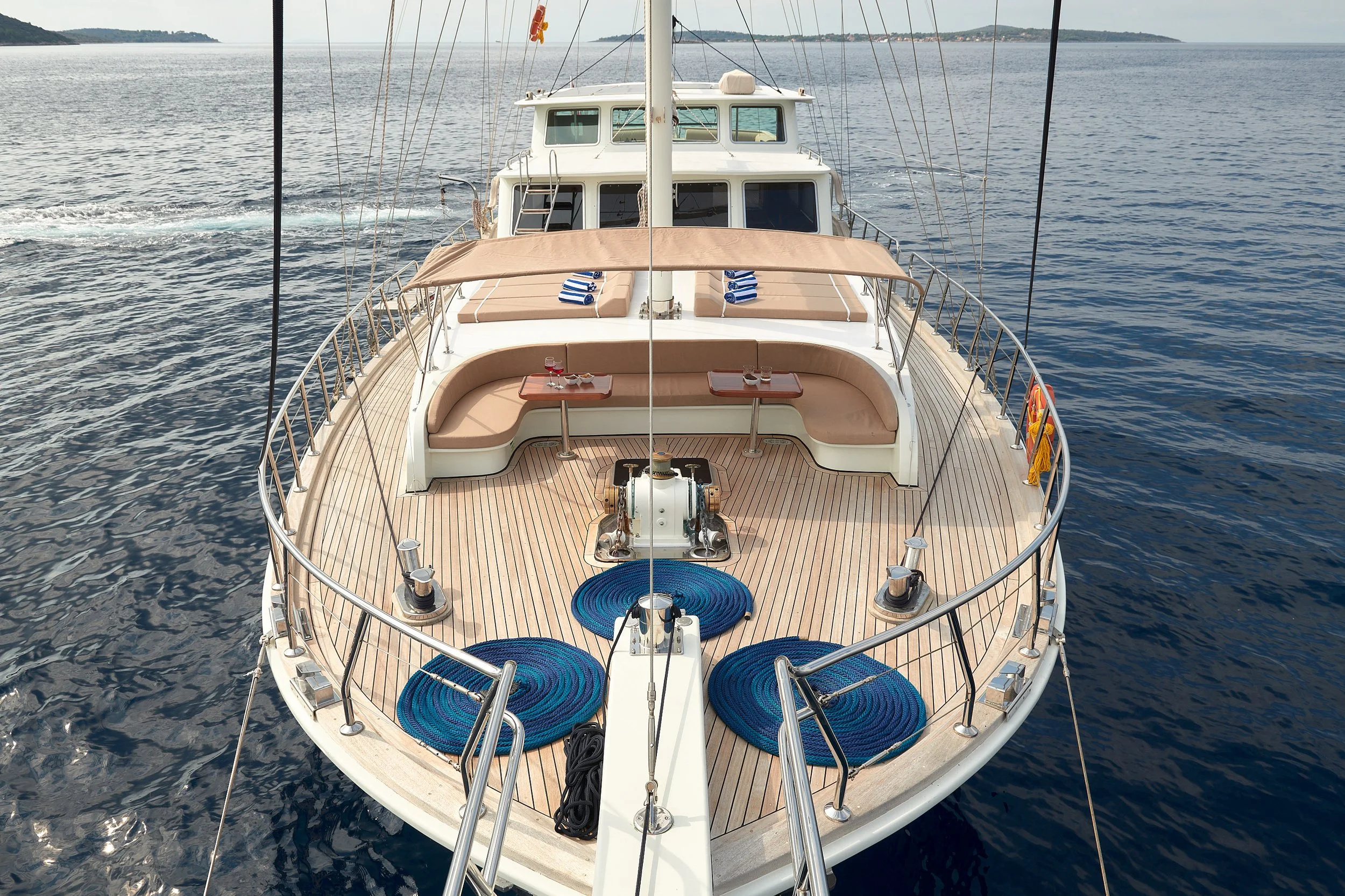 Front view of a yacht on the water with a spacious deck, beige cushioned seating, small tables, and blue rope coiled around two circular cushions. The ocean and a distant shoreline are visible in the background.