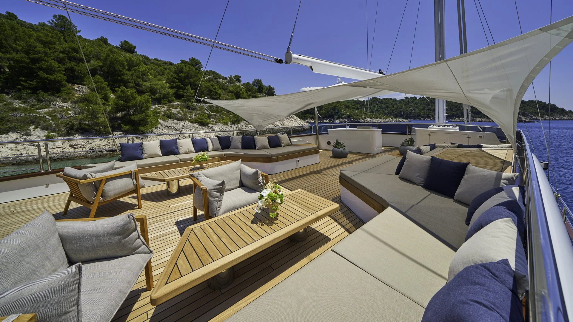 View of a luxury yacht deck with cushioned seating, wooden tables, and decorative plants, overlooking a body of water and a forested hillside under a clear blue sky.