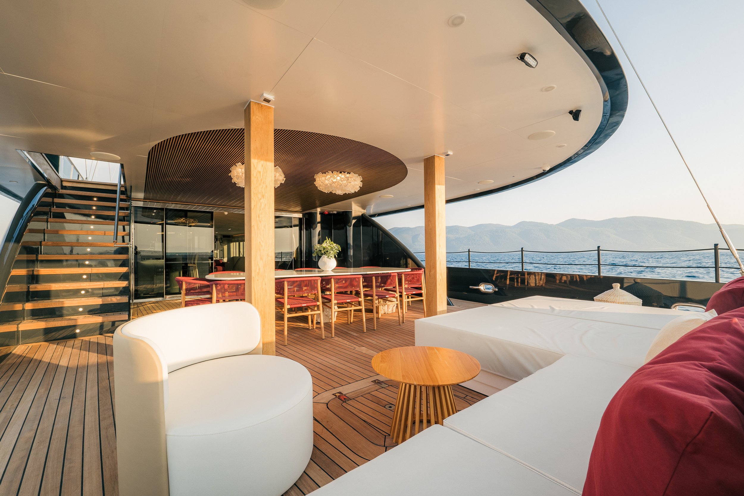Interior deck of a yacht showing seating areas, wooden flooring, staircase, and overlooking a body of water with mountains in the distance.