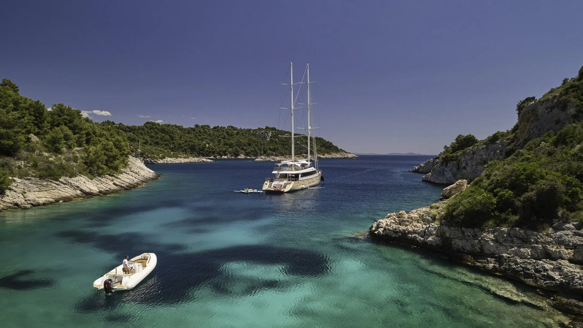 A large yacht with tall masts anchored between two lush, green, rocky islands with clear turquoise water surrounding it.