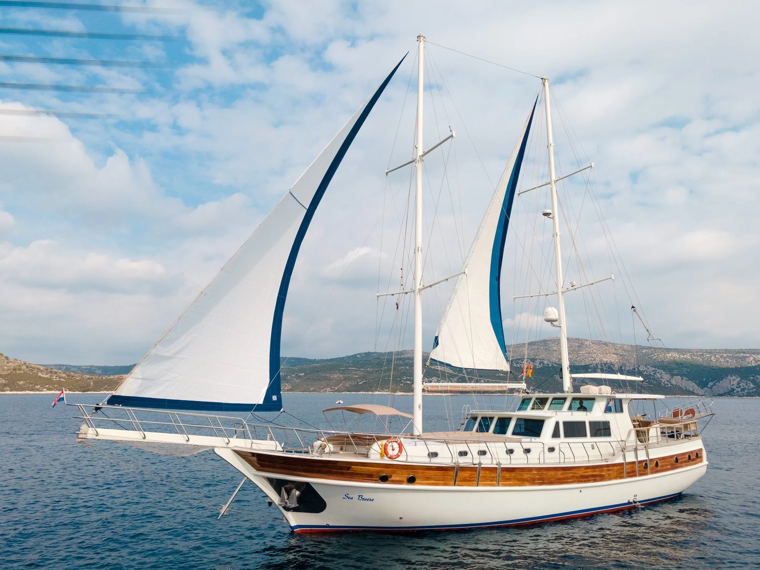 A sailboat with white and blue sails on calm water with a distant hilly shoreline under a partly cloudy sky.