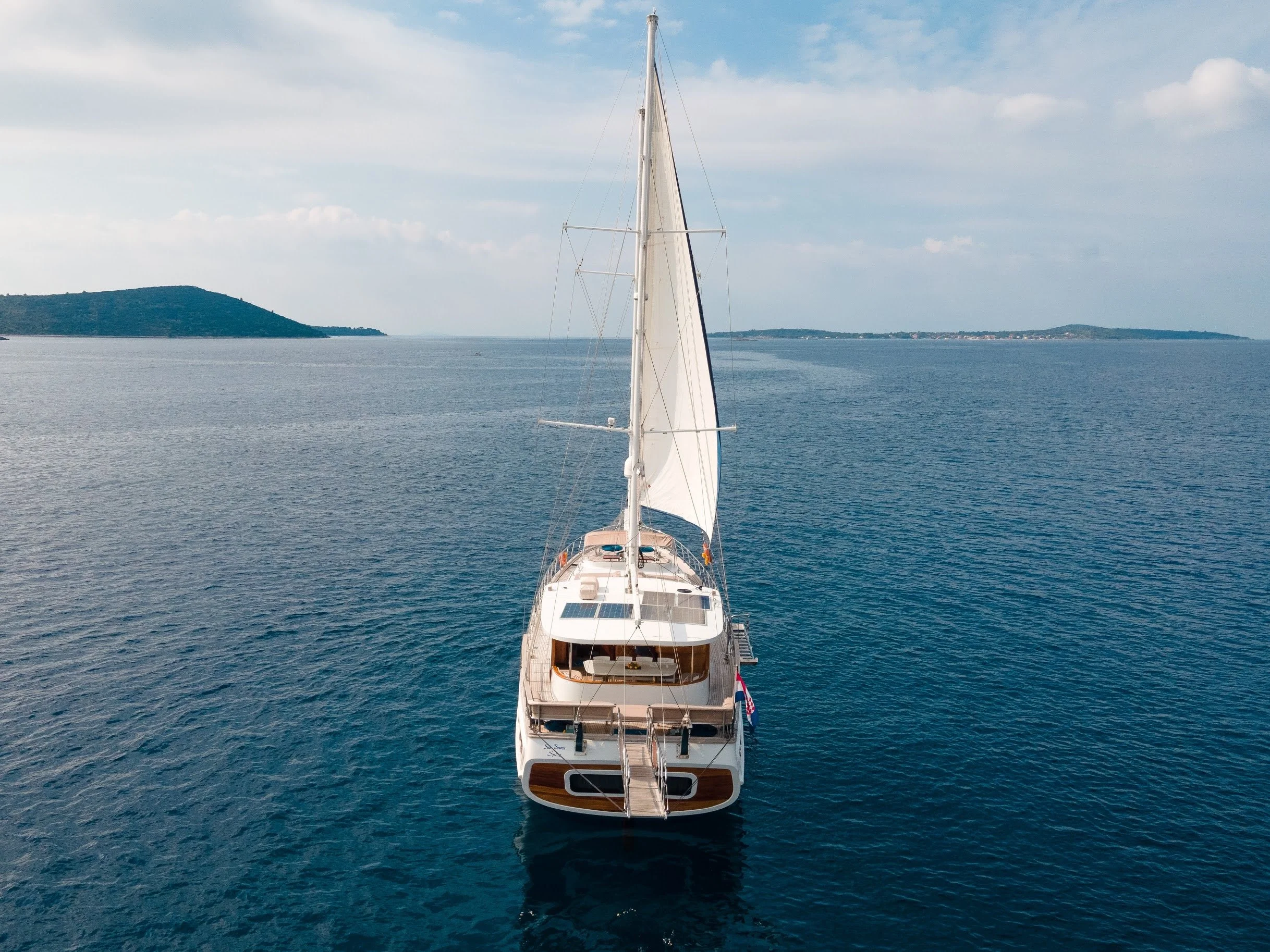 A sailboat afloat on calm blue waters with land in the distance and a partly cloudy sky overhead.