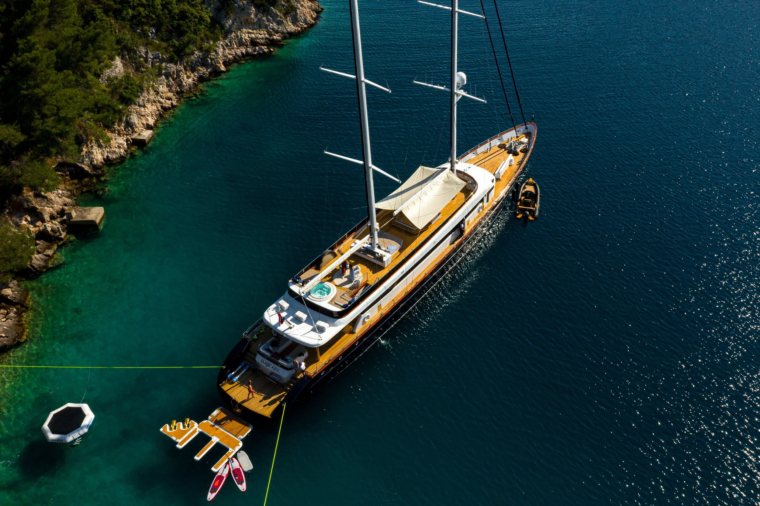 An aerial view of a large sailboat docked near a rocky shoreline with trees, in clear blue water. The boat has a hot tub, a shaded lounge area, and is accompanied by a small inflatable boat and paddleboards.
