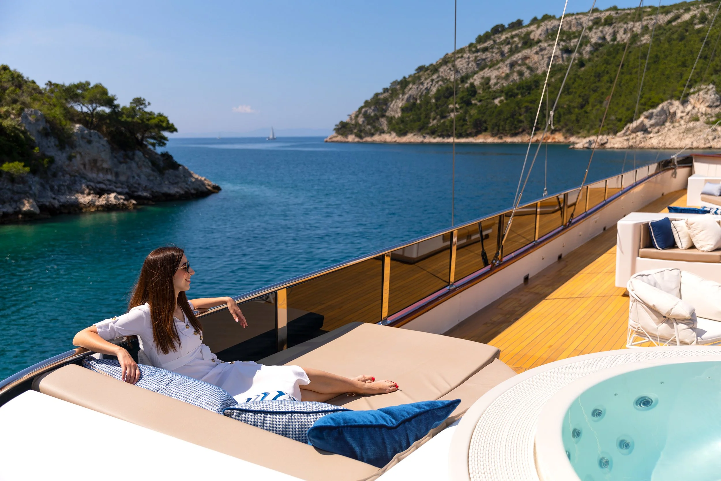 A woman sunbathing on a luxury yacht with a scenic view of the ocean, rocky islands, and sailboats in the distance under a clear blue sky.