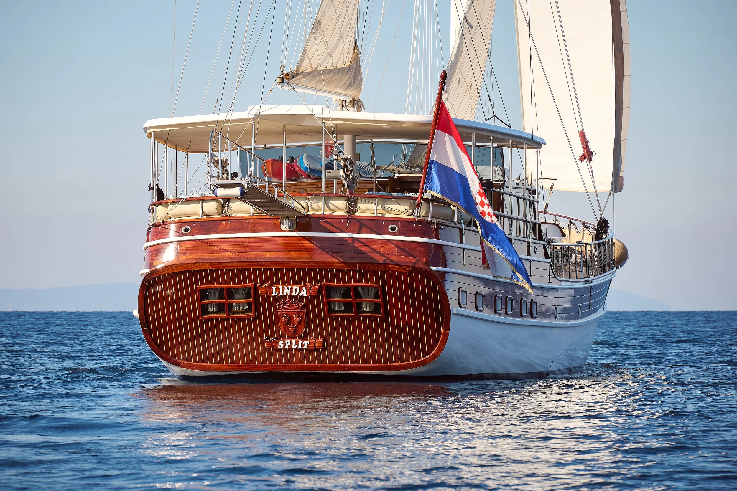 A large wooden sailing yacht with white sails and a Croatian flag on the stern, sailing on calm blue water with a distant horizon in the background.