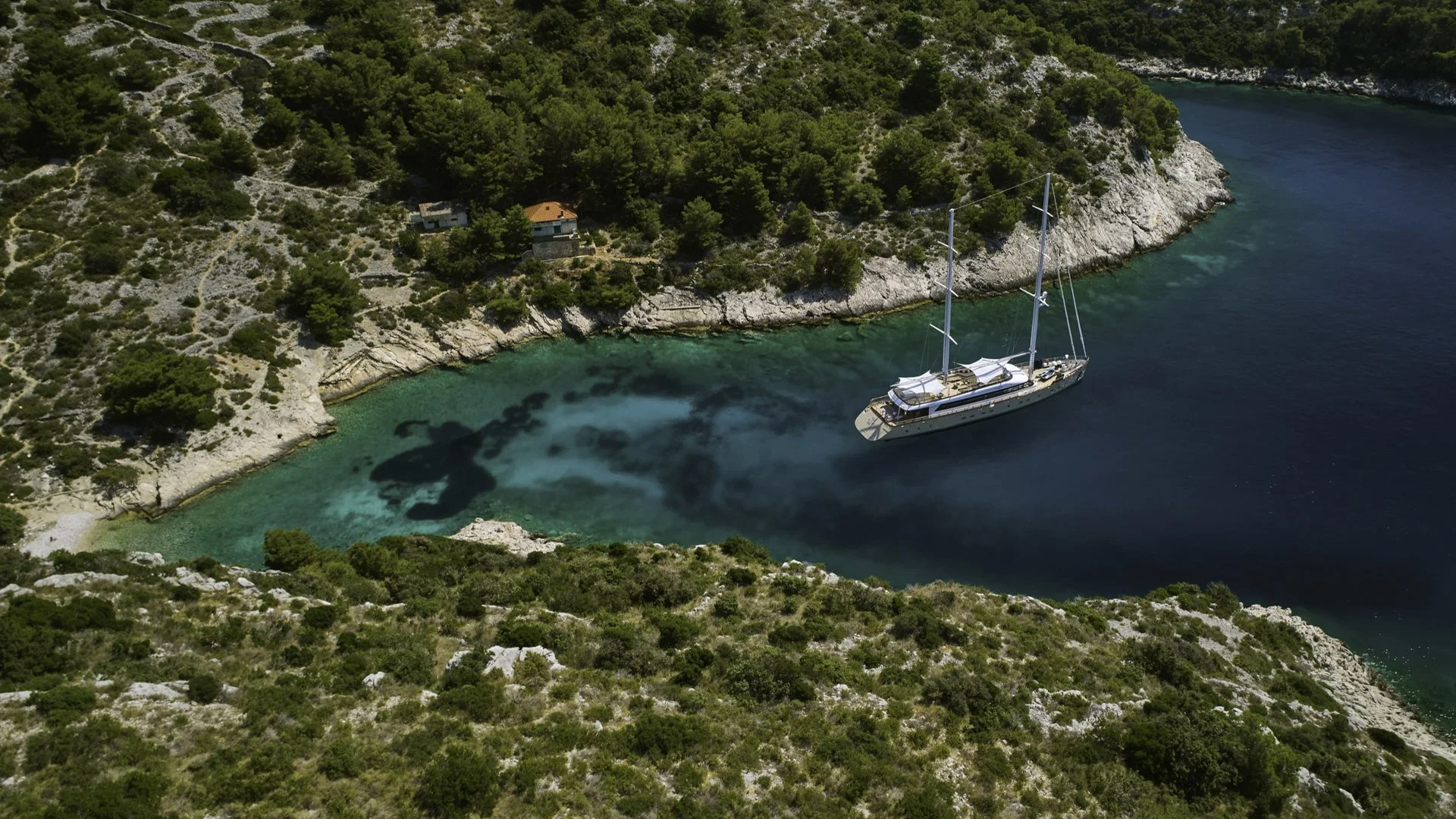An aerial view of a sailboat anchored in a small, clear, turquoise cove surrounded by rocky, green hillside with vegetation and trees.