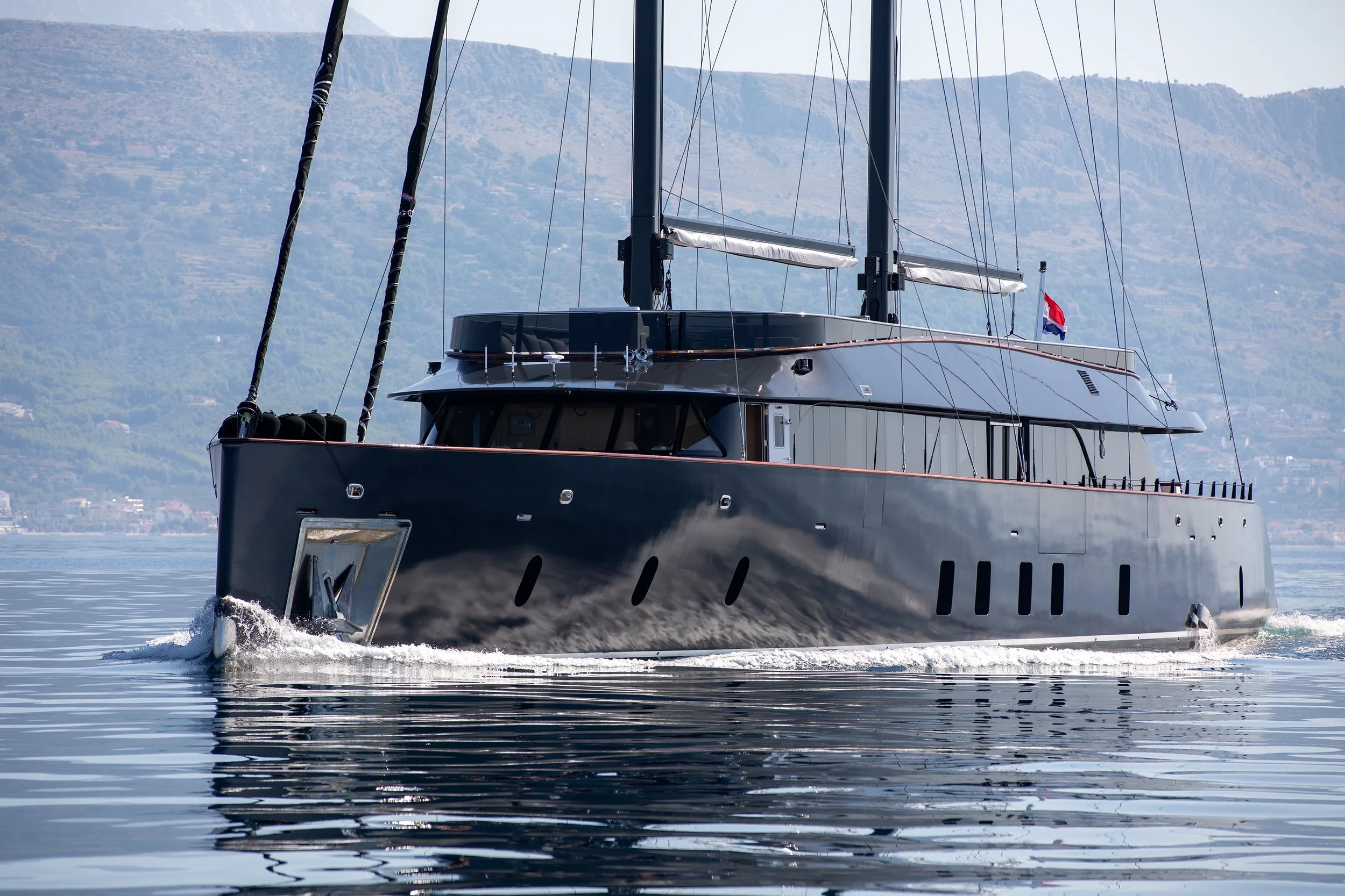A sleek black sailing yacht with two masts sailing on calm water, with a mountainous landscape in the background.