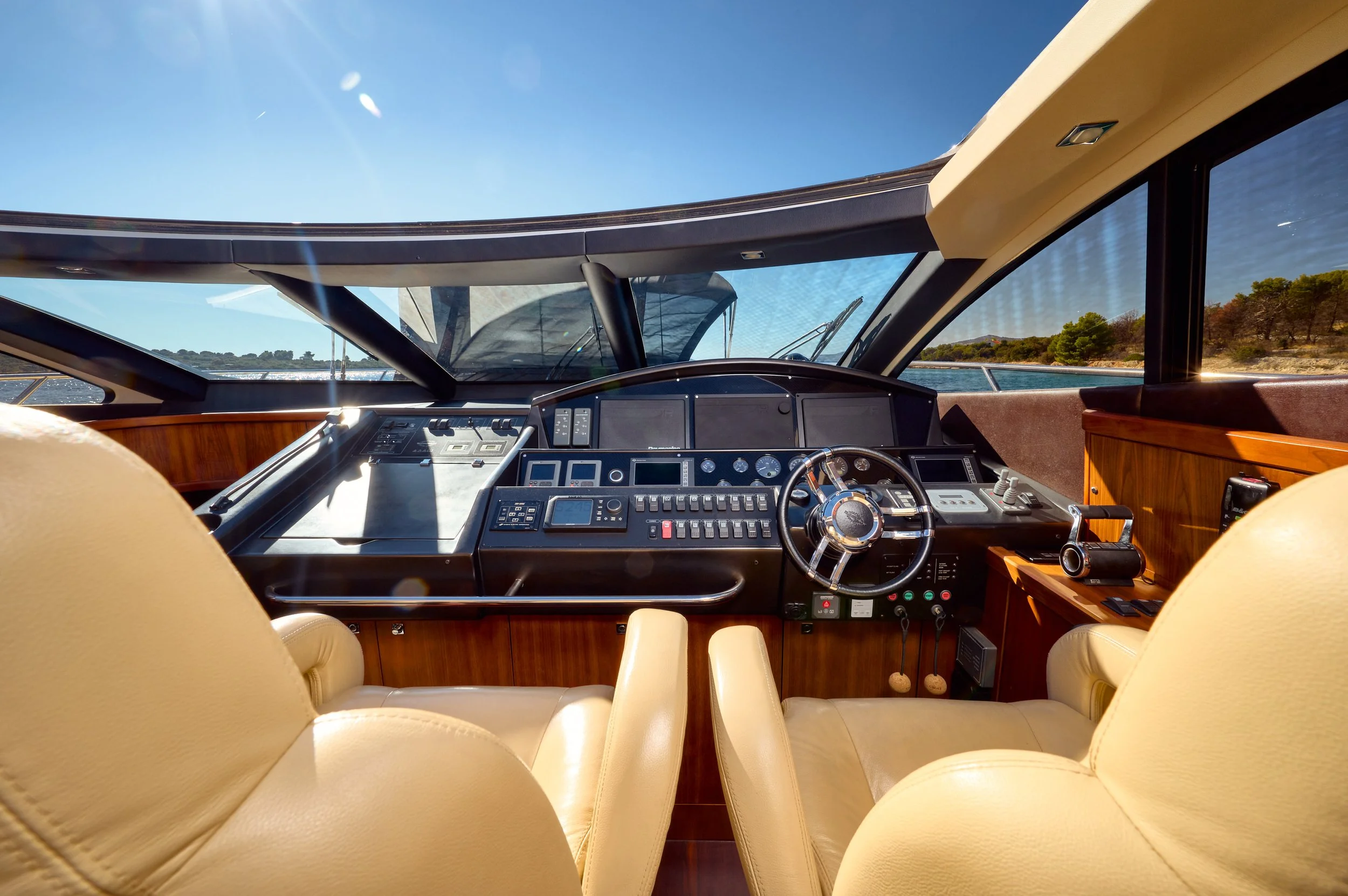 View of a yacht's cockpit with a steering wheel, various navigation instruments, control panels, and two cream-colored leather seats, with water and trees visible through the large front windows.
