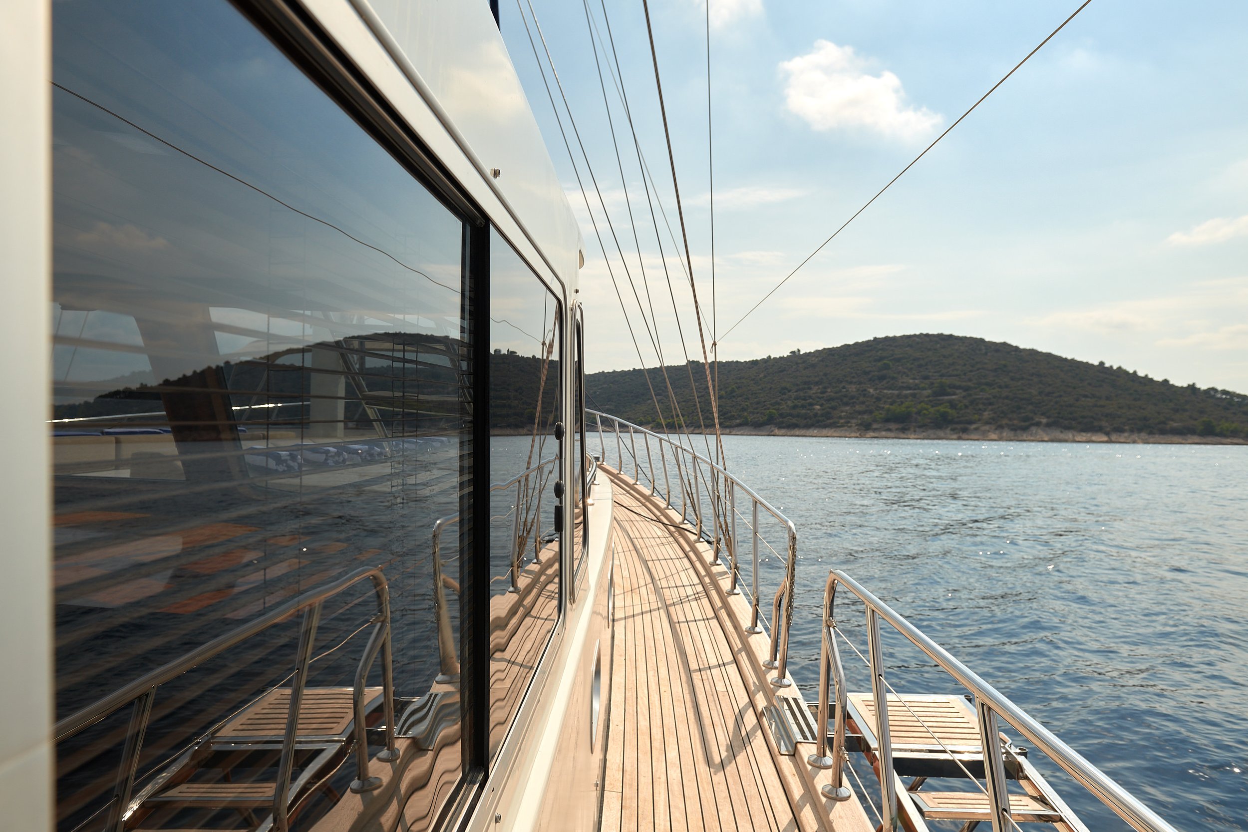 View from a boat with a wooden deck and metal railings, looking forward towards a distant landmass with hills, water, and a partly cloudy sky.