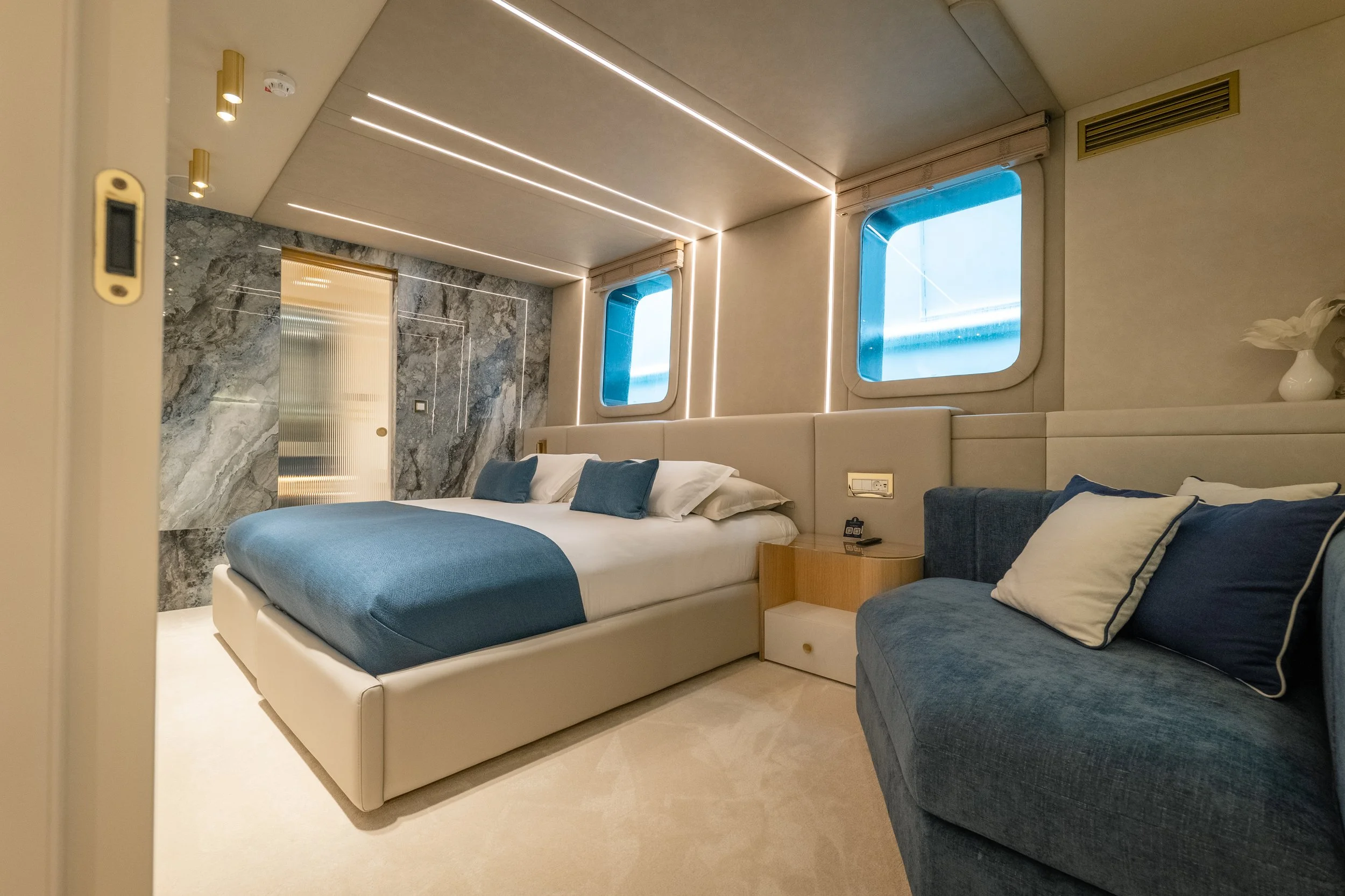A modern hotel bedroom with a large bed, blue and white bedding, and two pillows. There's a small blue sofa with pillows, a wooden nightstand, and two windows with blue sky visible outside. The room has a minimalist design with marble accents and war