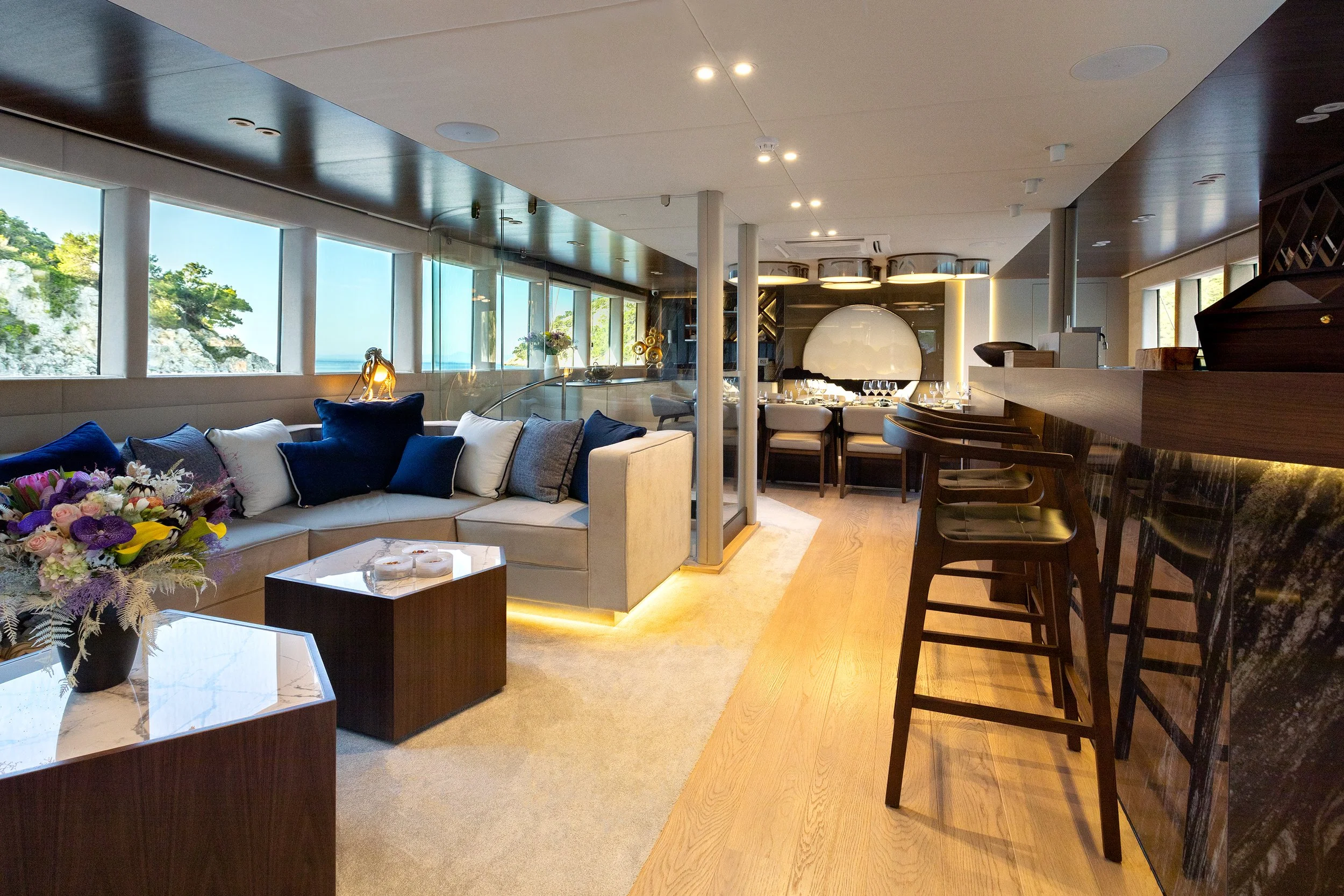 Interior of a modern living space with a view of the ocean, featuring a beige sectional sofa with colorful pillows, flowers, wooden coffee tables, a bar area with barstools, and large windows showing a scenic coastal landscape.
