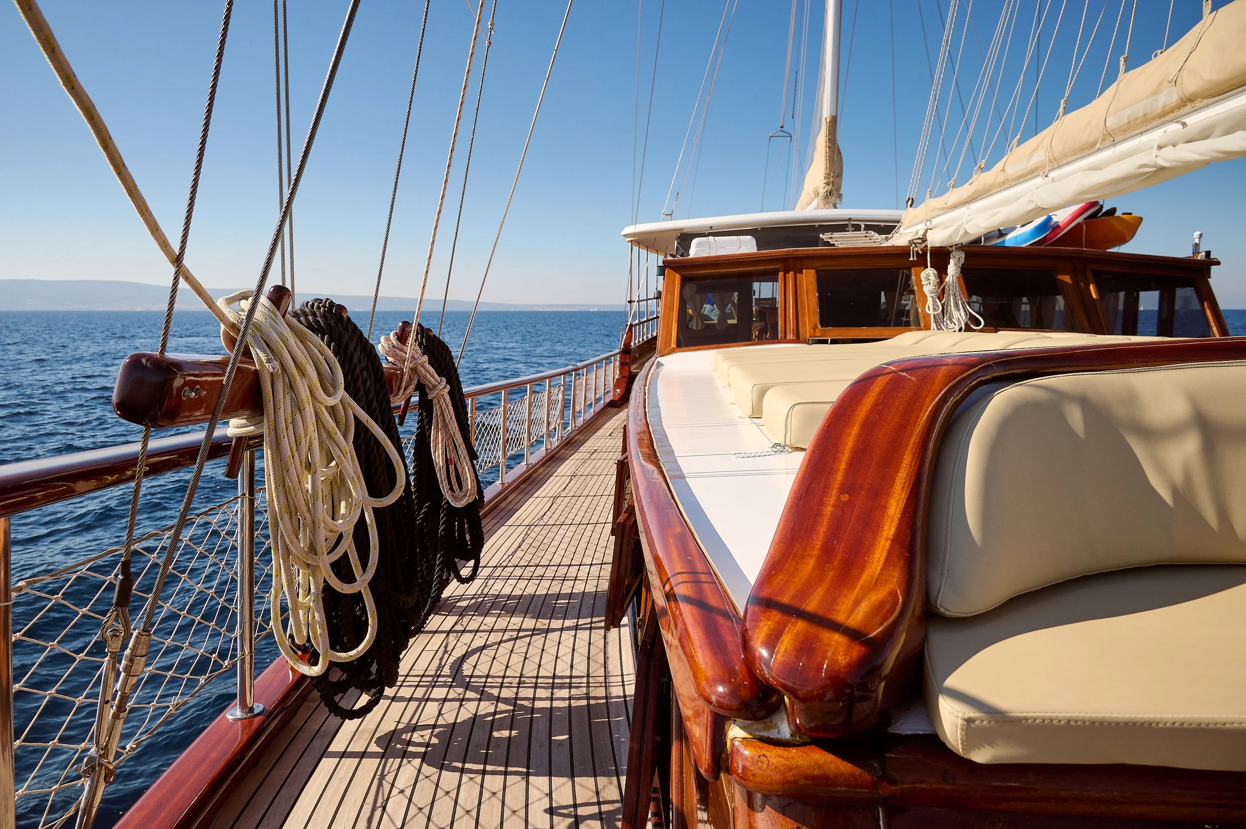 A view from a sailboat deck with coiled ropes hanging on a wooden railing, glass windshields, and cushioned seating, overlooking the open ocean on a clear sunny day.