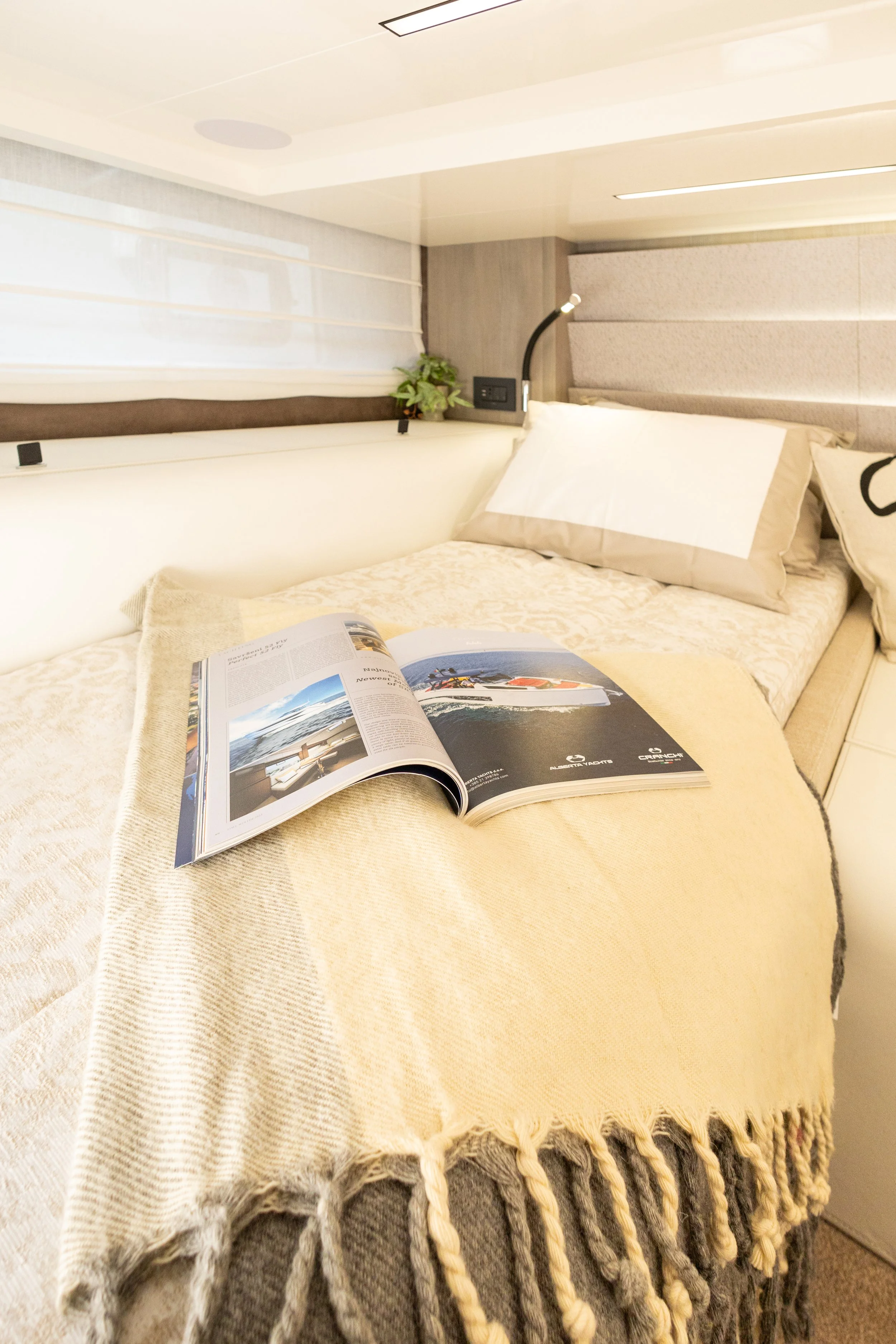 A neatly made bed with a beige blanket and pillows, an open magazine on top, a small plant and reading lamp attached to the headboard, in a well-lit room with a window on the left.