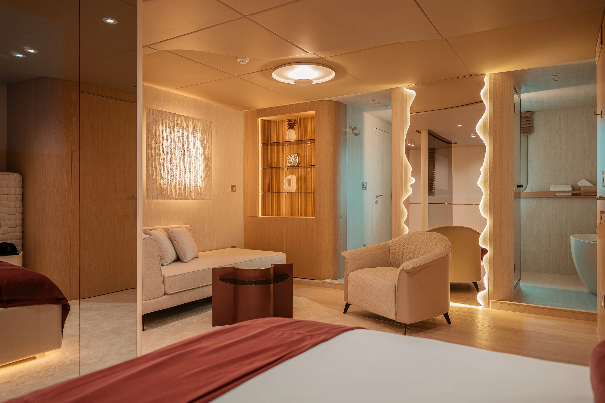 Interior of a modern hotel room with warm lighting, a beige sofa, a cream armchair, a small dark wood coffee table, a bed with burgundy bedding, a built-in wooden shelf with decor, a textured wall art, and an open bathroom with a shower and sink.