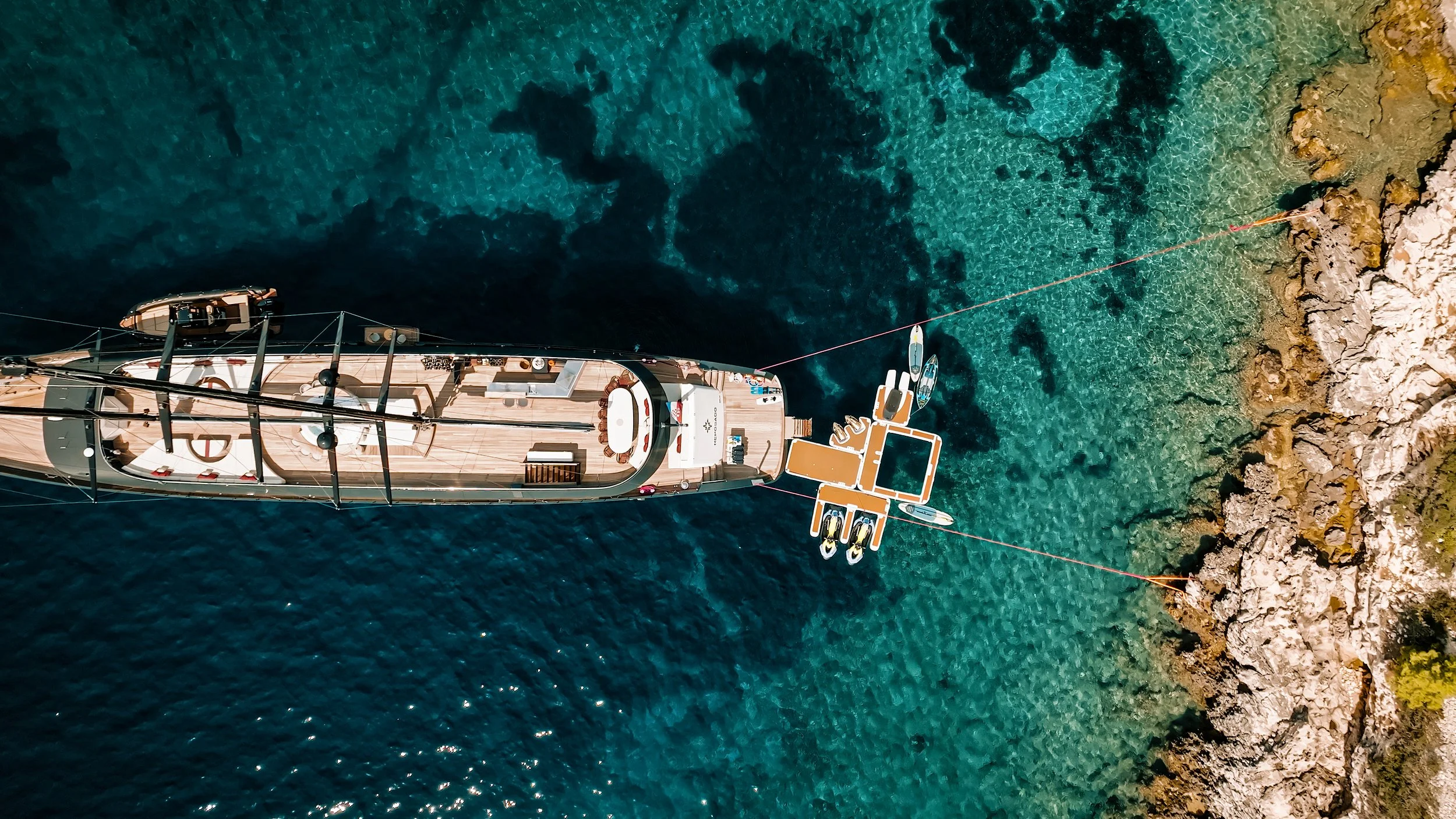 An aerial view of a large yacht anchored near a rocky shoreline with clear turquoise water, and a smaller boat attached to the yacht's floating platform.