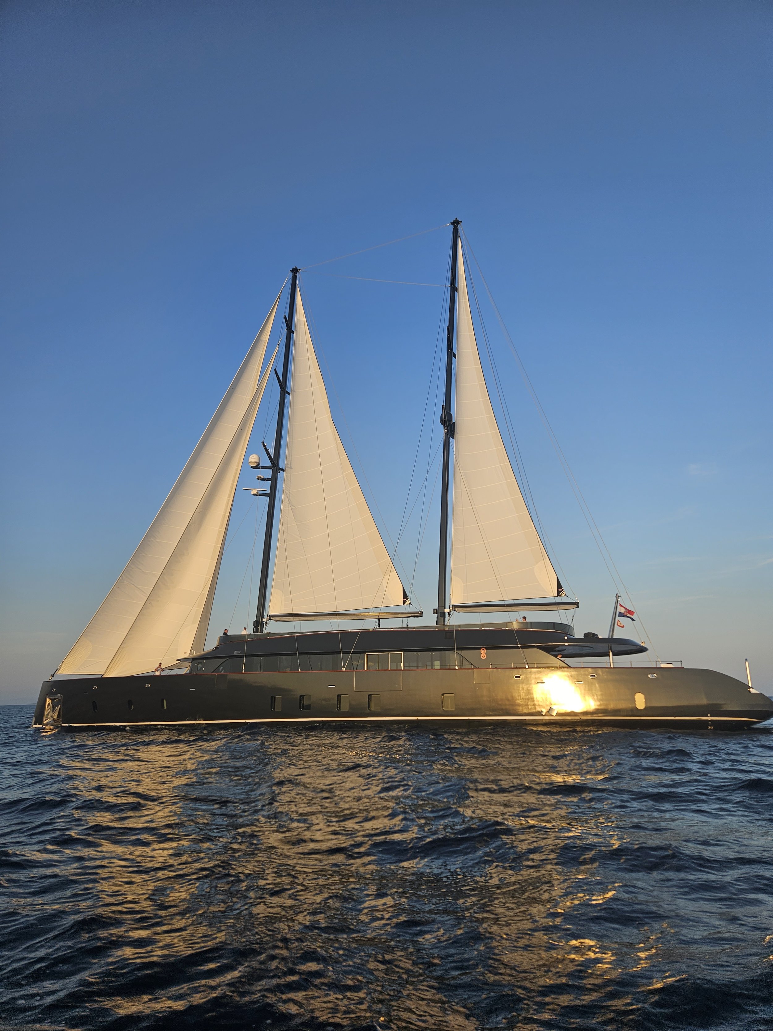 A large black sailing yacht with white sails on the ocean during sunset.