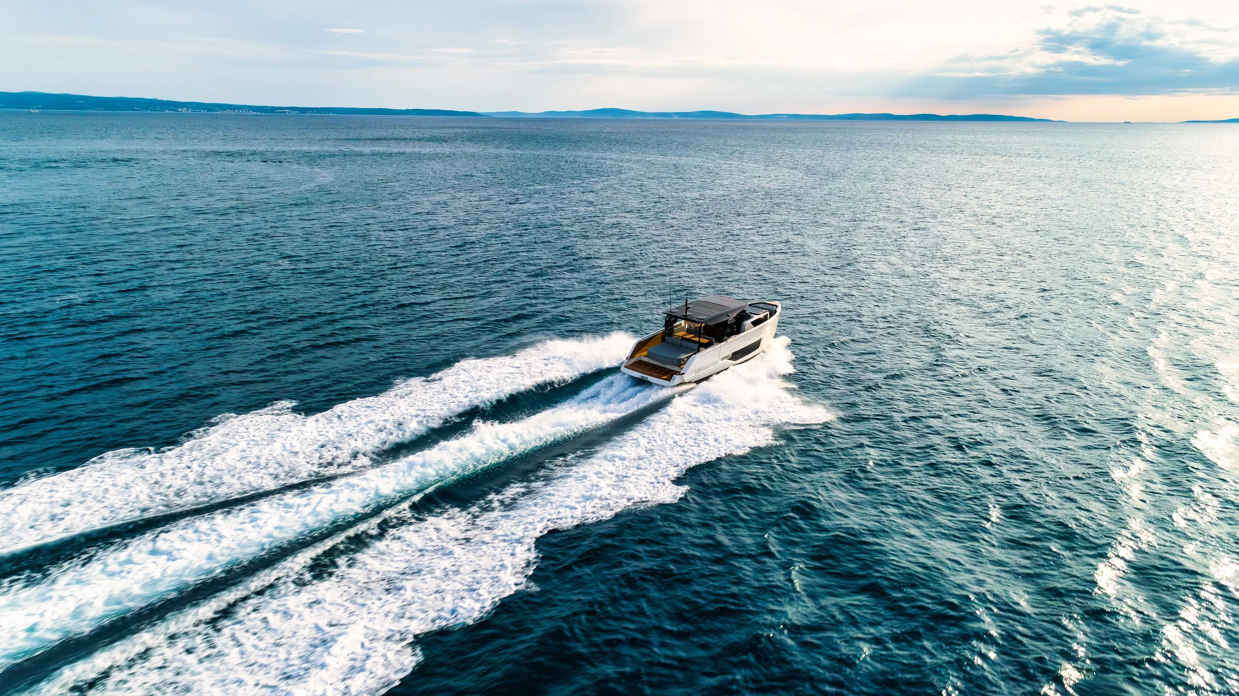 A yacht cruising on the ocean creating a white wake behind it with a distant shoreline under a partly cloudy sky.